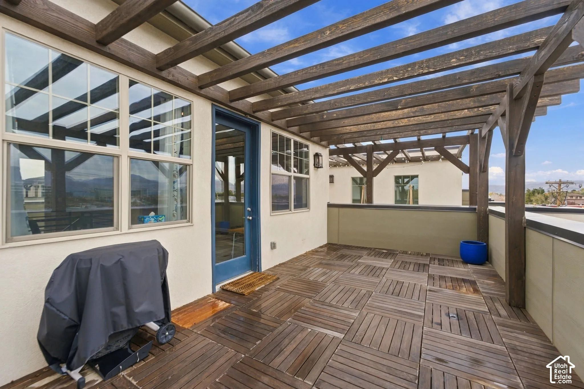 Rooftop deck featuring a timber pergola, modular wood-finish patio tiles, stucco walls, and a glass-paneled entry door