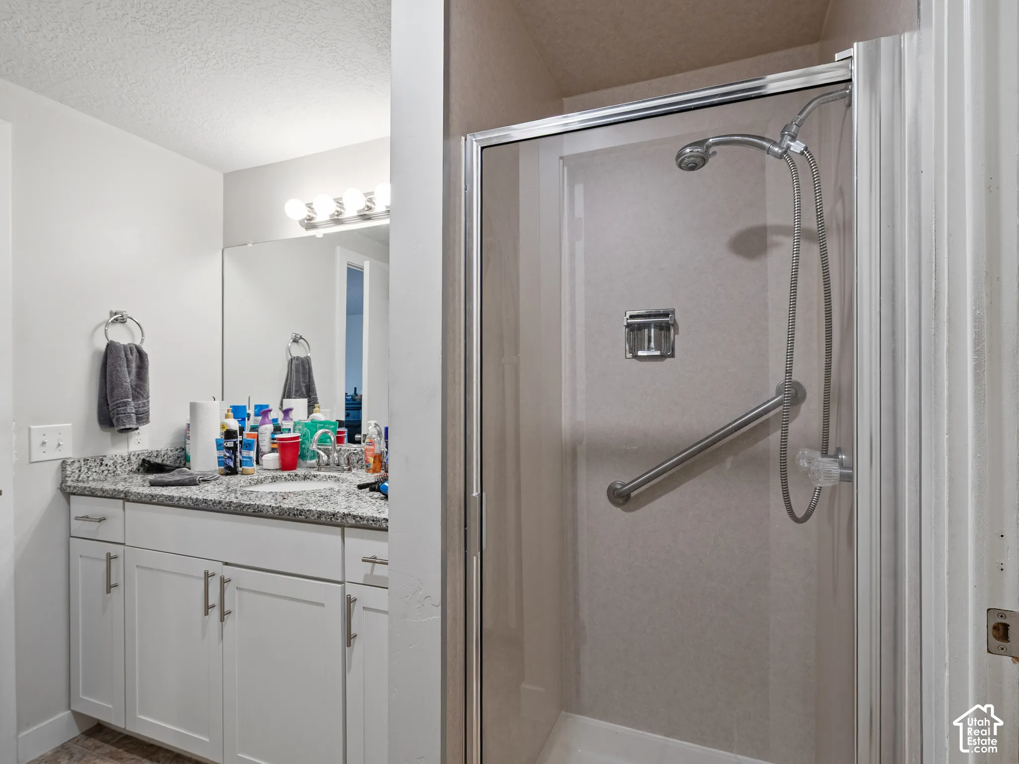 Bathroom featuring white shaker-style cabinetry, stone-surface countertop, full-width vanity mirror, and glass-enclosed shower with stainless steel hardware