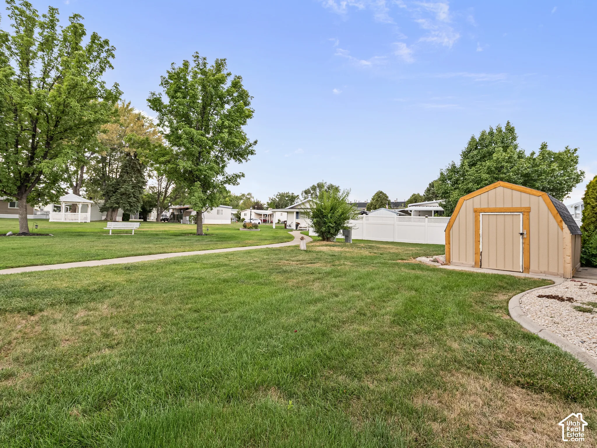 Expansive yard featuring a manicured lawn, detached storage shed, white vinyl perimeter fencing, concrete walkway, and mature trees