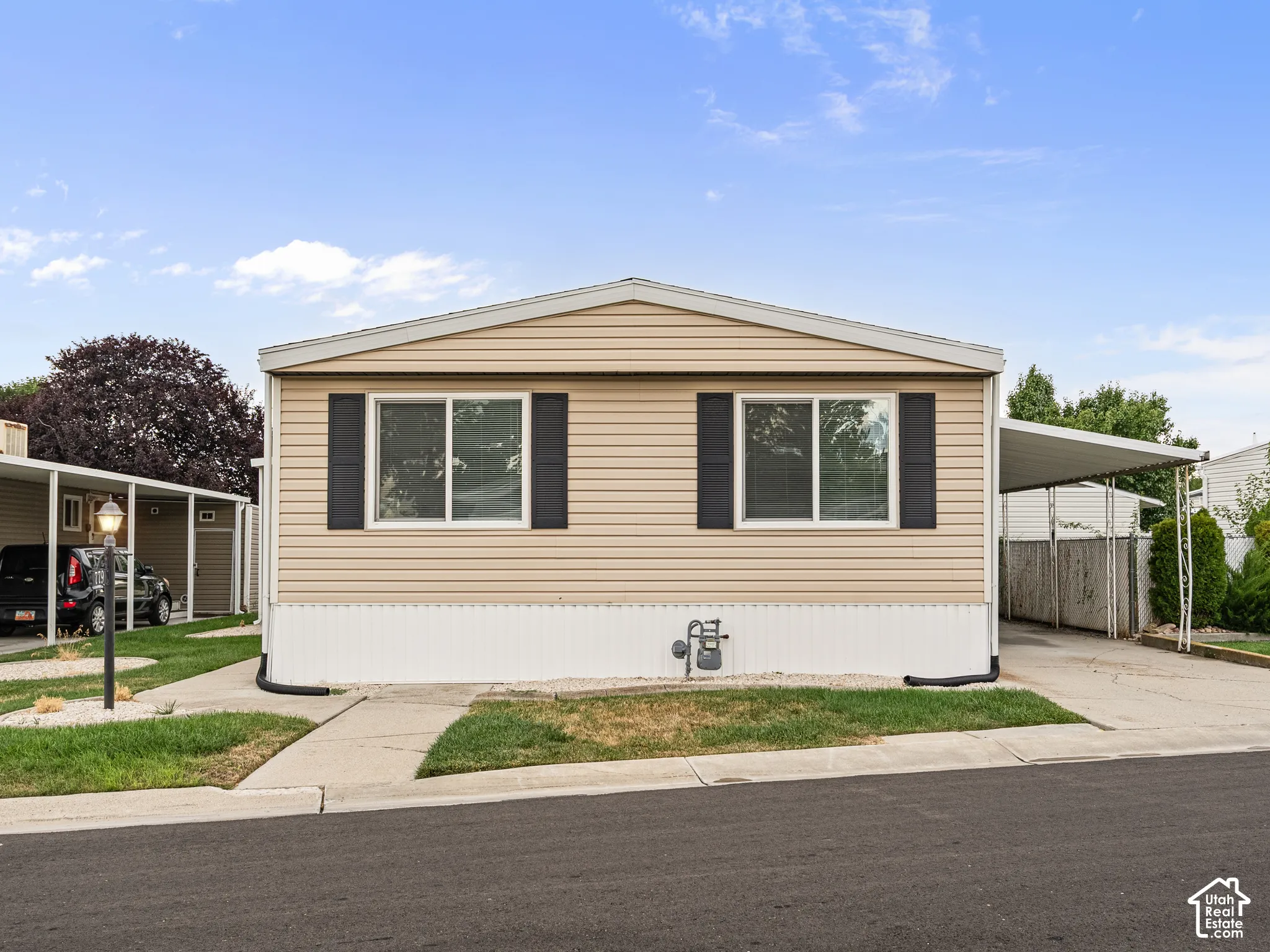 Tan vinyl siding exterior featuring black window shutters, a white foundation skirting, and an attached carport