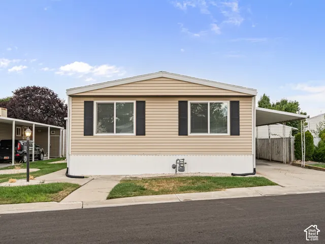Tan vinyl siding exterior featuring black window shutters, a white foundation skirting, and an attached carport