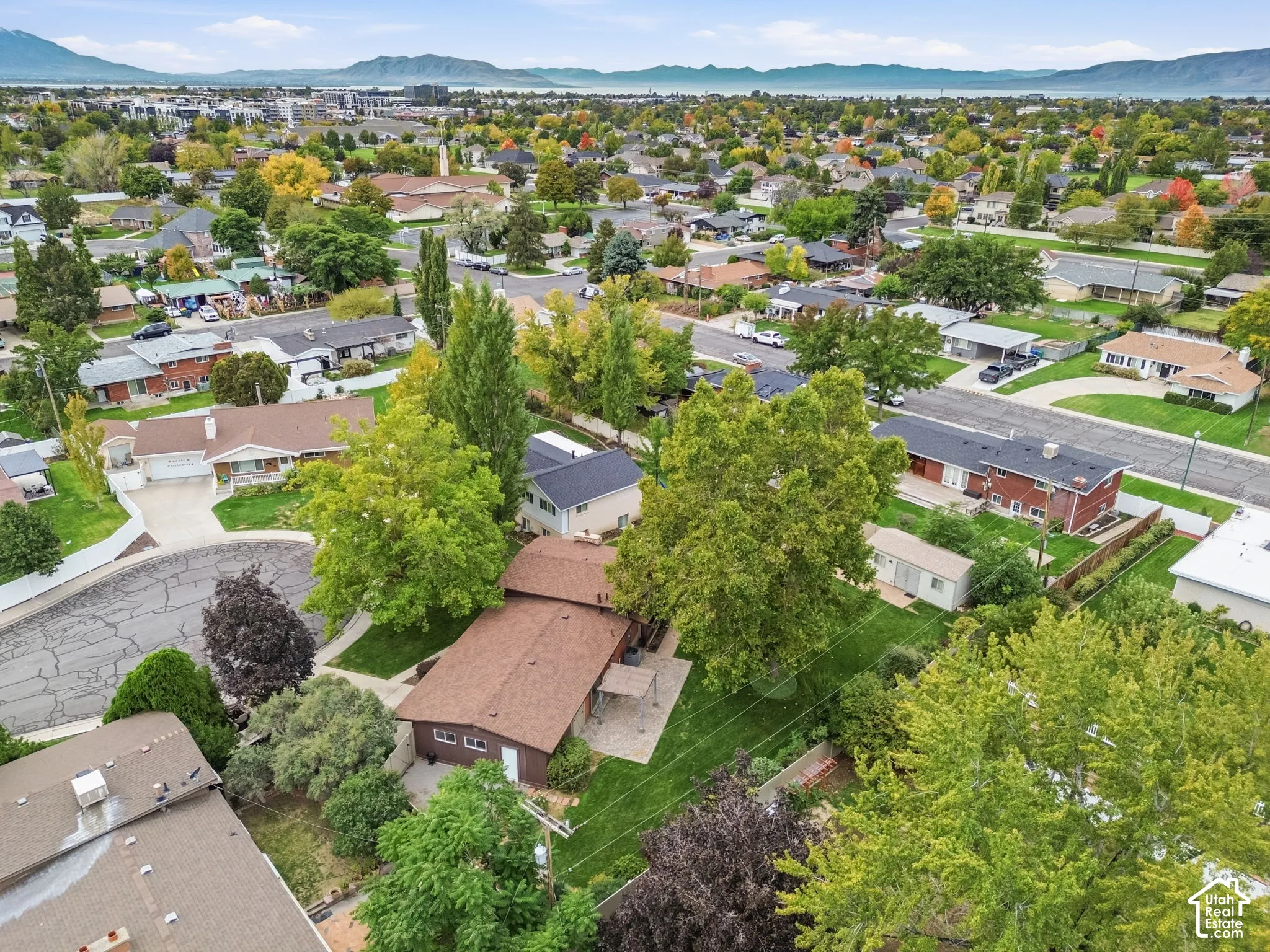 Aerial perspective featuring an expansive lot with mature trees, a paved patio area, and a backdrop of mountain scenery