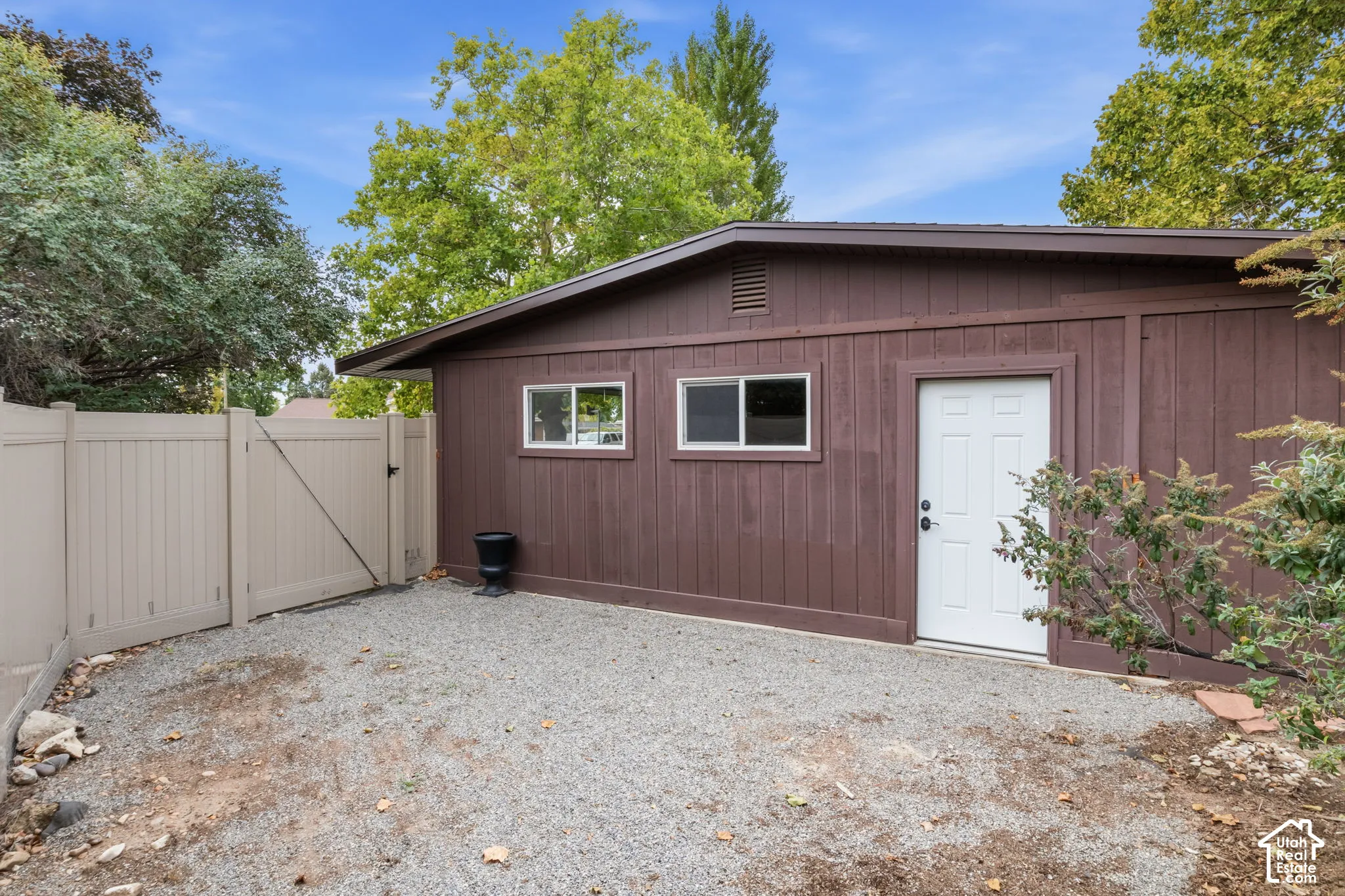 Detached structure featuring wood-panel siding, dual sliding windows, an exterior entry door, and a gravel yard area enclosed by vinyl privacy fencing