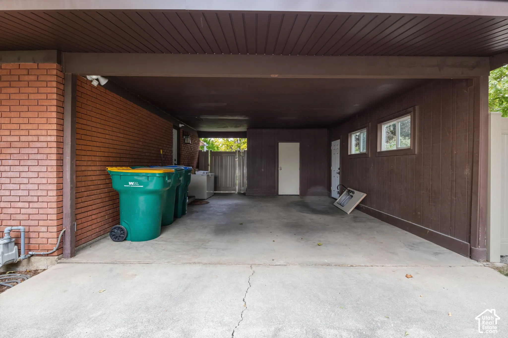 Covered carport featuring a concrete floor, red brick exterior wall, dark wood-finish siding, and two exterior access doors