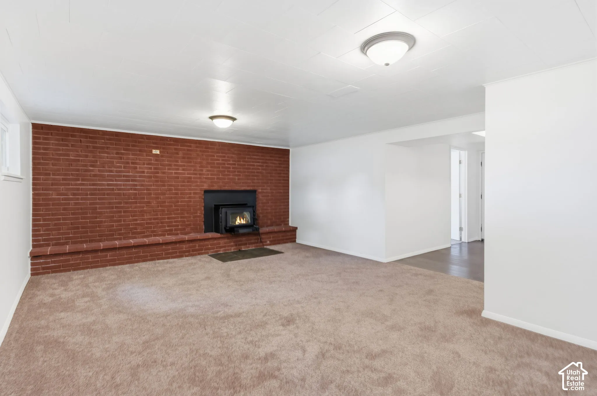 Expansive room featuring a floor-to-ceiling red brick accent wall, a built-in fireplace insert with a brick hearth, neutral wall-to-wall carpeting, and dual flush-mount ceiling light fixtures