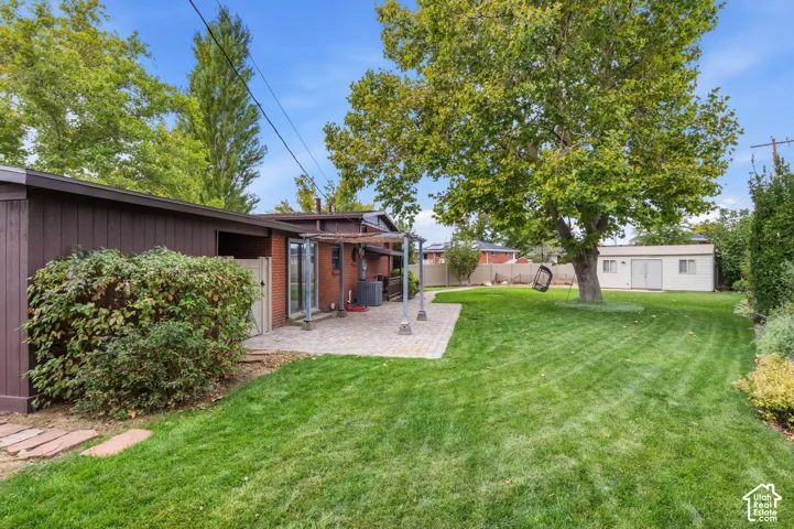Backyard featuring a paved stone patio, mature trees, lawn, detached storage structure, and privacy fencing