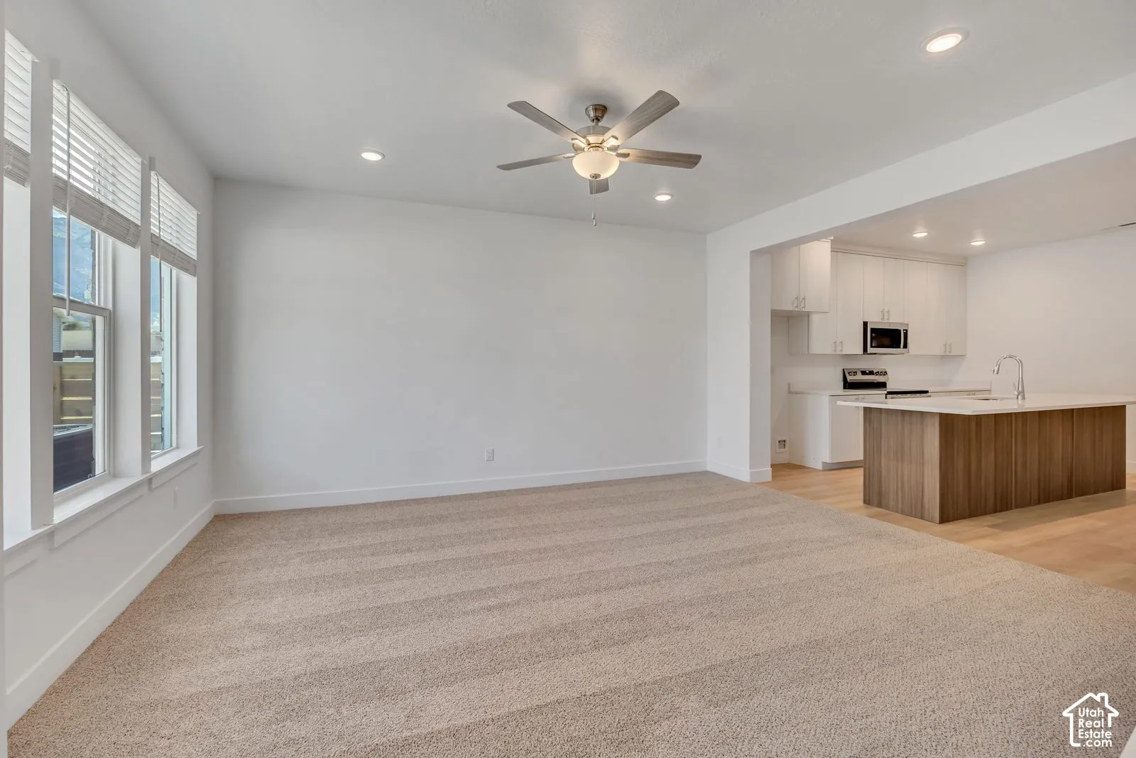 Neutral-toned carpeting, recessed ceiling lighting, a central ceiling fan with integrated light fixture, large windows with horizontal blinds, and a kitchen featuring white cabinetry and a wood-finish island