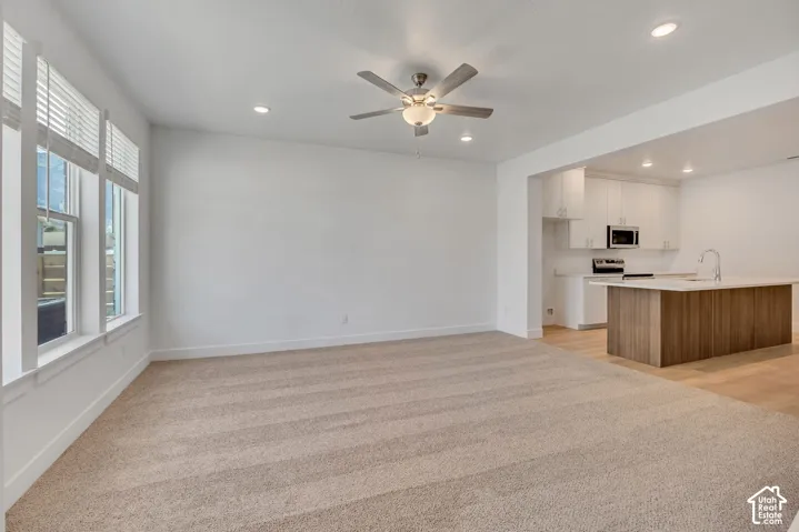 Neutral-toned carpeting, recessed ceiling lighting, a central ceiling fan with integrated light fixture, large windows with horizontal blinds, and a kitchen featuring white cabinetry and a wood-finish island