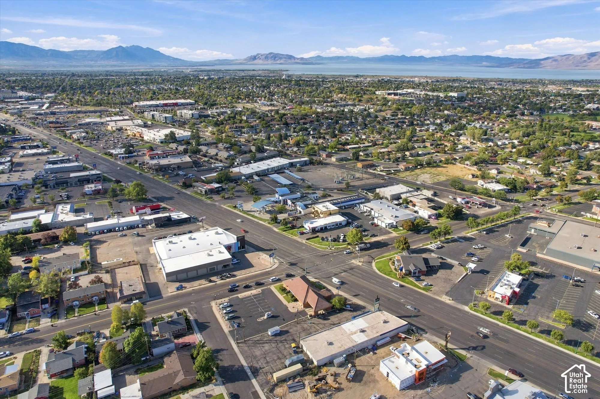 Aerial perspective of a commercial district featuring paved roadway infrastructure, multi-tenant building complexes, asphalt parking surfaces, and mountain range scenery