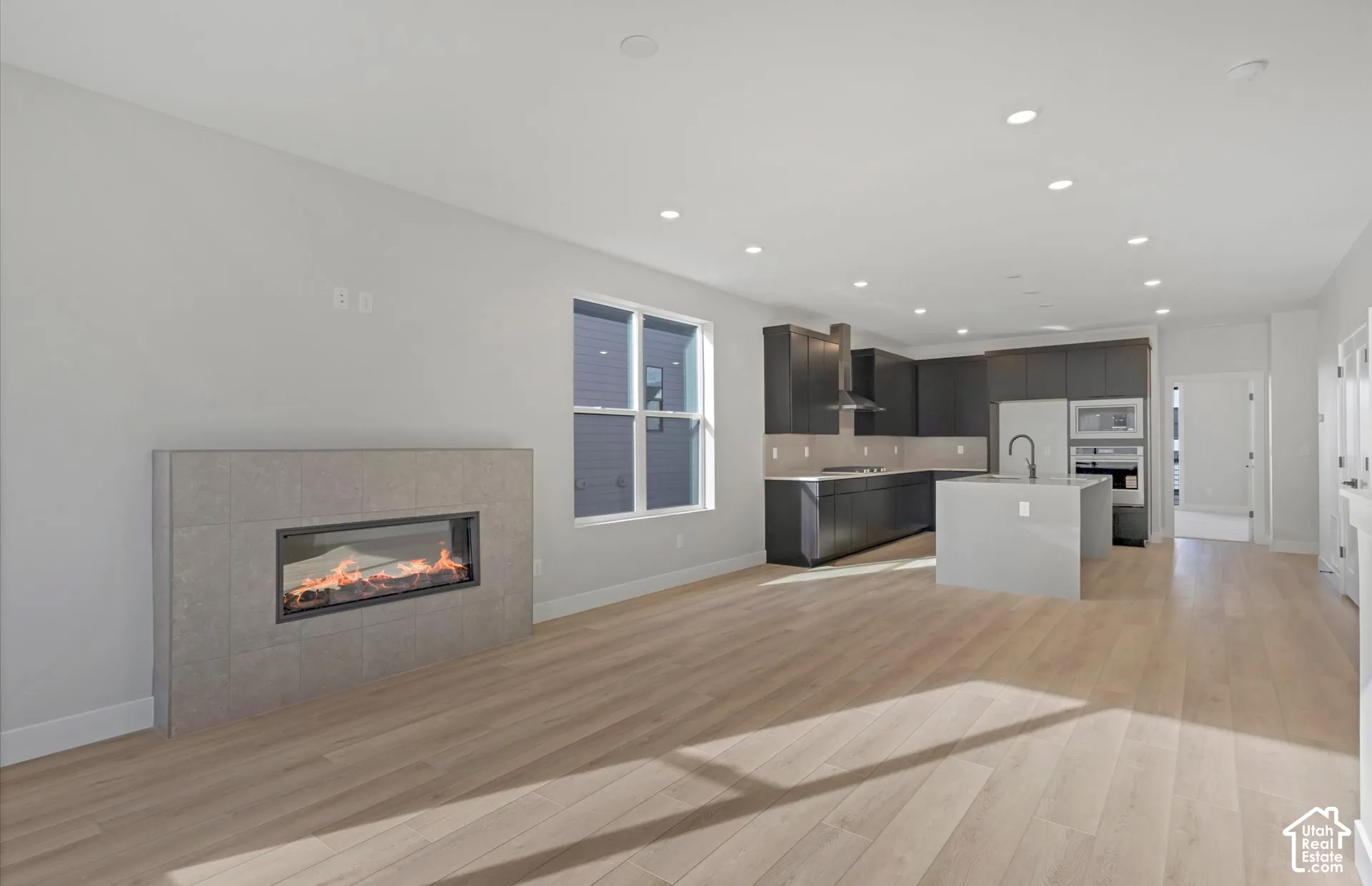 Linear fireplace with a tiled surround, wood-finish flooring, recessed lighting, central kitchen island with countertop, and dark-toned cabinetry