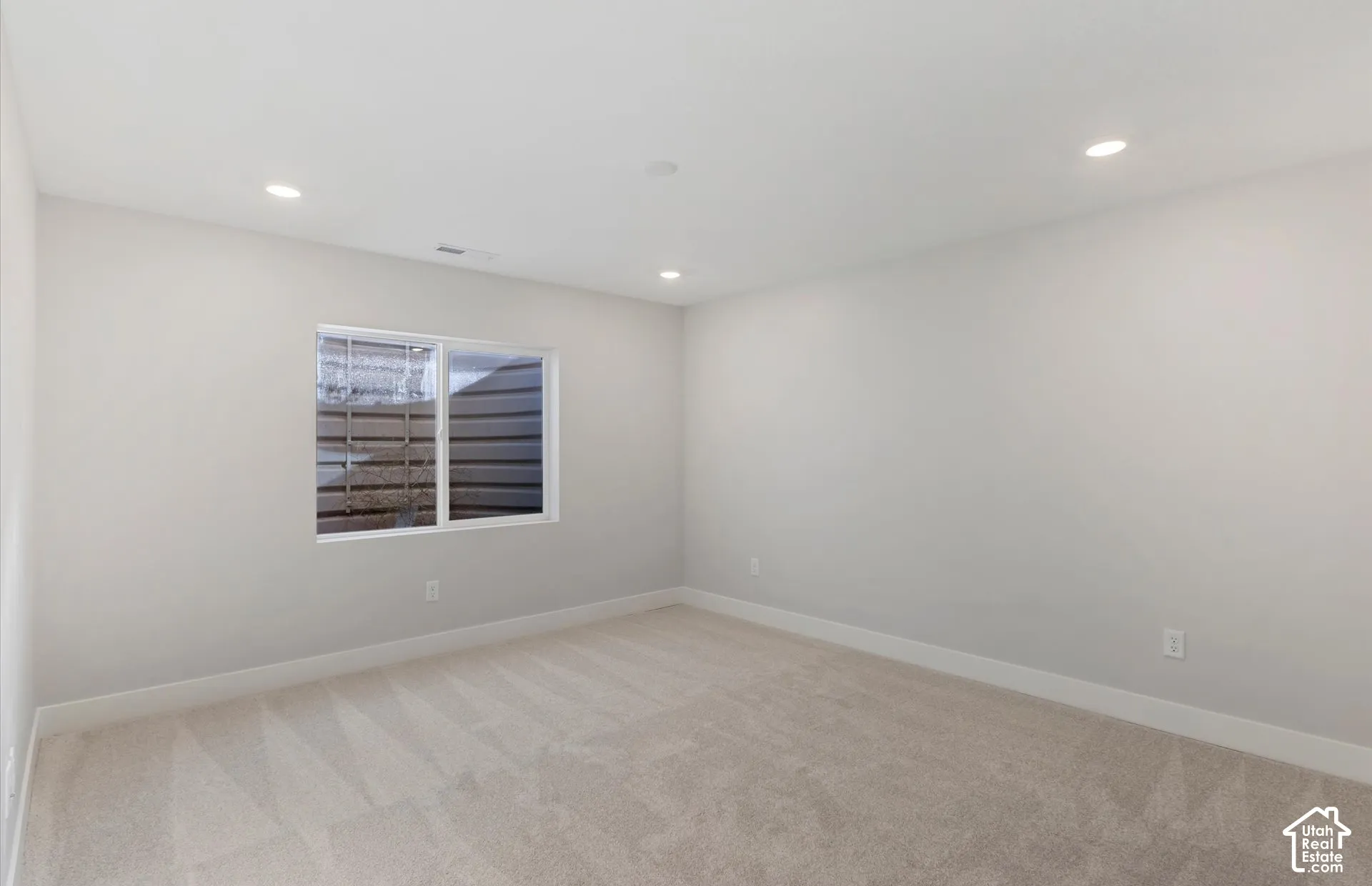 Neutral-toned room featuring carpeted flooring, recessed lighting, and a window with a corrugated metal window well