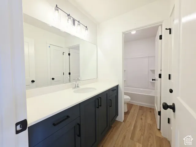 Dark-toned shaker cabinetry with matte black hardware, white countertop with an integrated basin, full-width wall mirror, linear vanity lighting, and wood-finish flooring