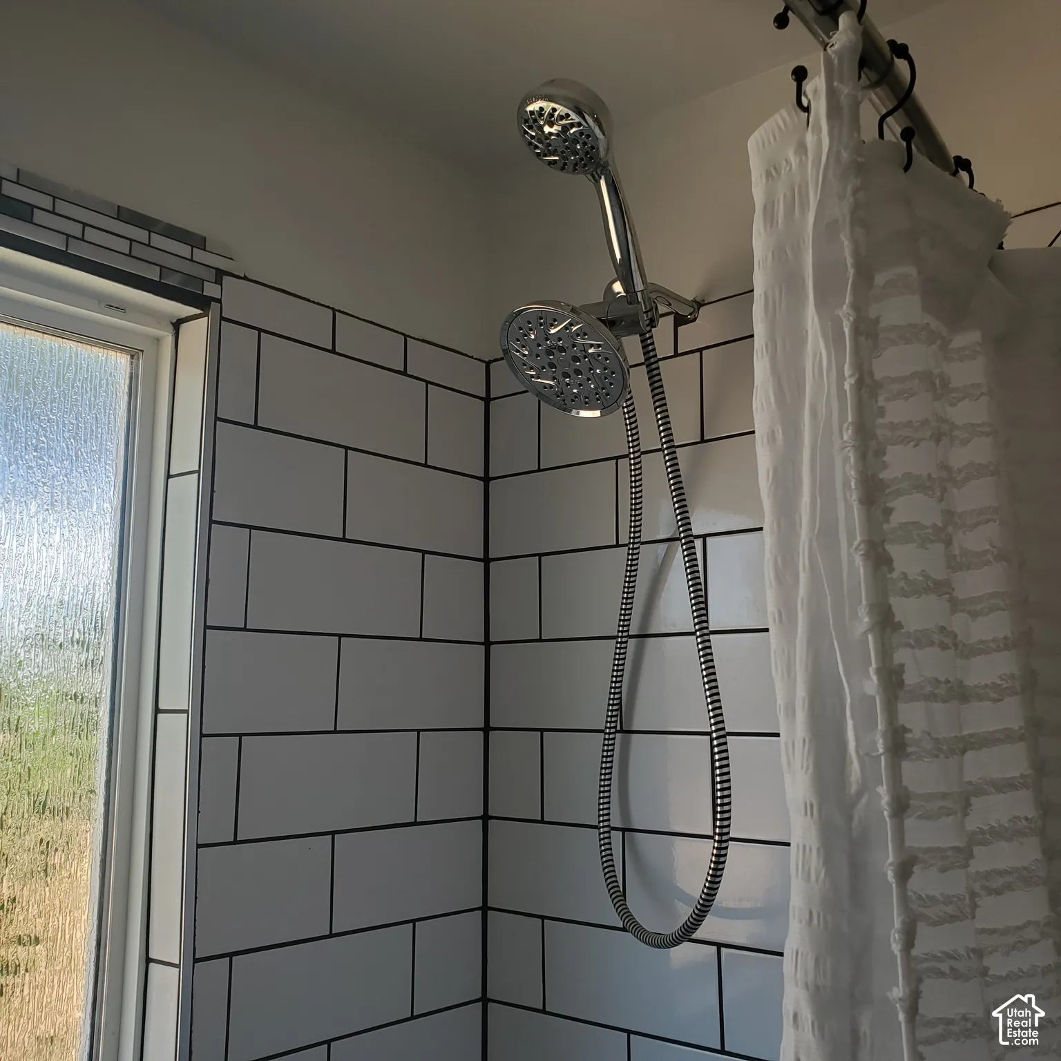 Shower enclosure featuring white rectangular tile with dark grout, chrome dual-head shower fixture, and window with textured privacy glass