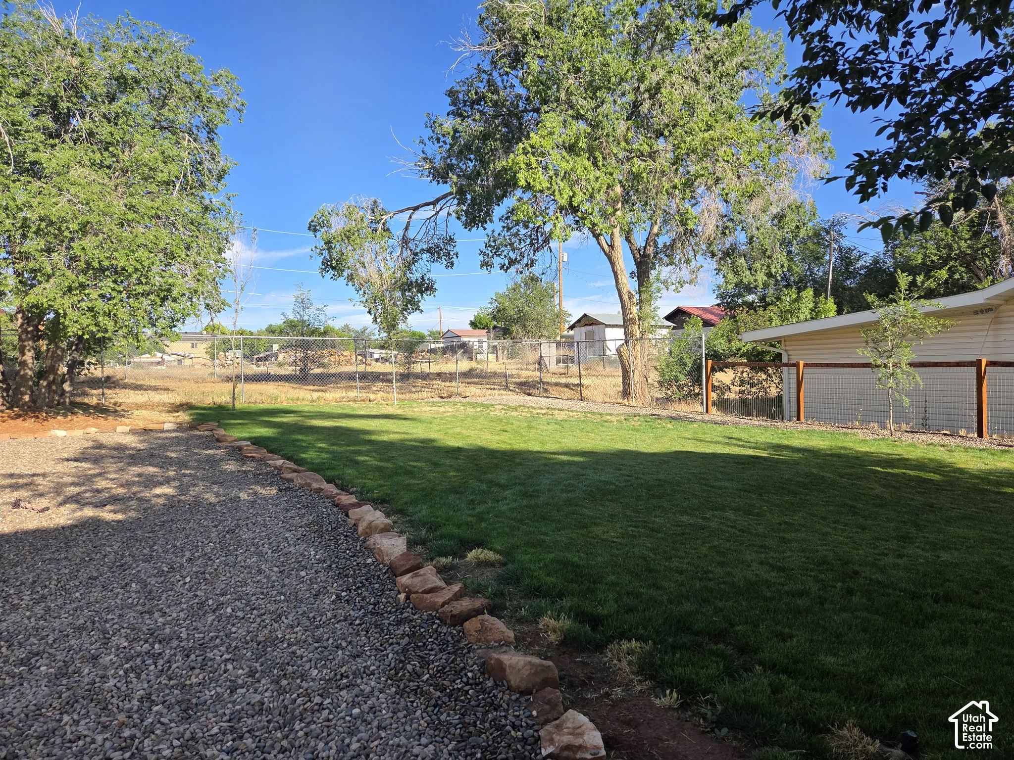 Backyard featuring a manicured lawn, stone border edging, gravel ground cover, mature trees, and perimeter fencing.