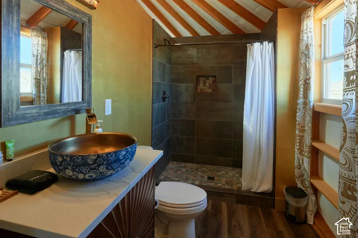 Bathroom featuring a vessel sink with patterned finish, stone-tile shower surround with integrated niche, wood-finish flooring, and exposed ceiling rafters