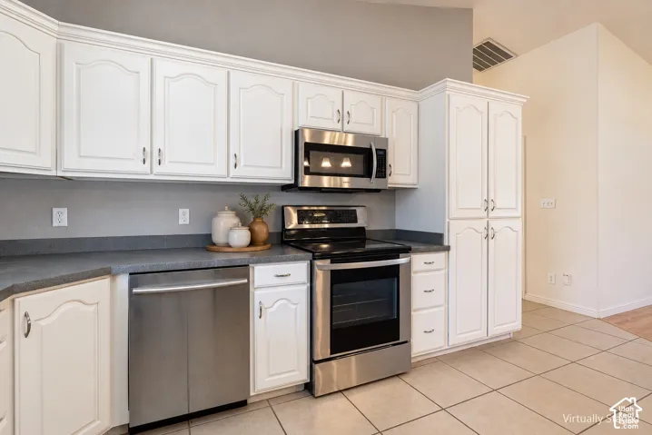 Kitchen featuring white cabinetry, stainless steel appliances, dark solid-surface countertops, and tiled flooring under a vaulted ceiling