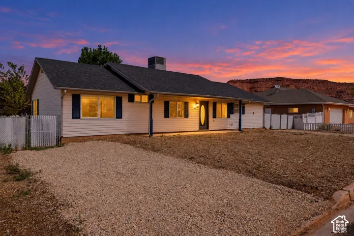 Single-level exterior featuring light-toned horizontal siding, dark decorative shutters, a gravel driveway, and a shingle roof set against a prominent red rock formation backdrop