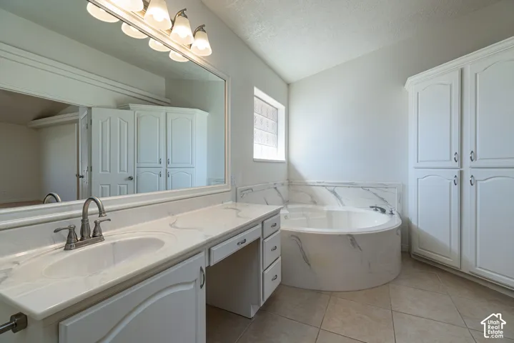 Bathroom featuring a vanity with integrated sink and stone-patterned countertop, a matching stone-patterned surround soaking tub, full-width mirror, built-in floor-to-ceiling cabinetry, and tile flooring