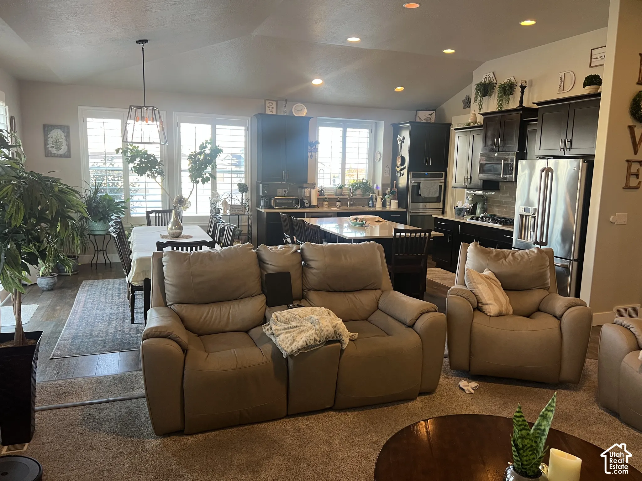 Open-concept kitchen featuring dark-toned cabinetry, a central island with stone-surface countertop, stainless steel appliances, recessed ceiling lighting, and windows fitted with plantation shutters