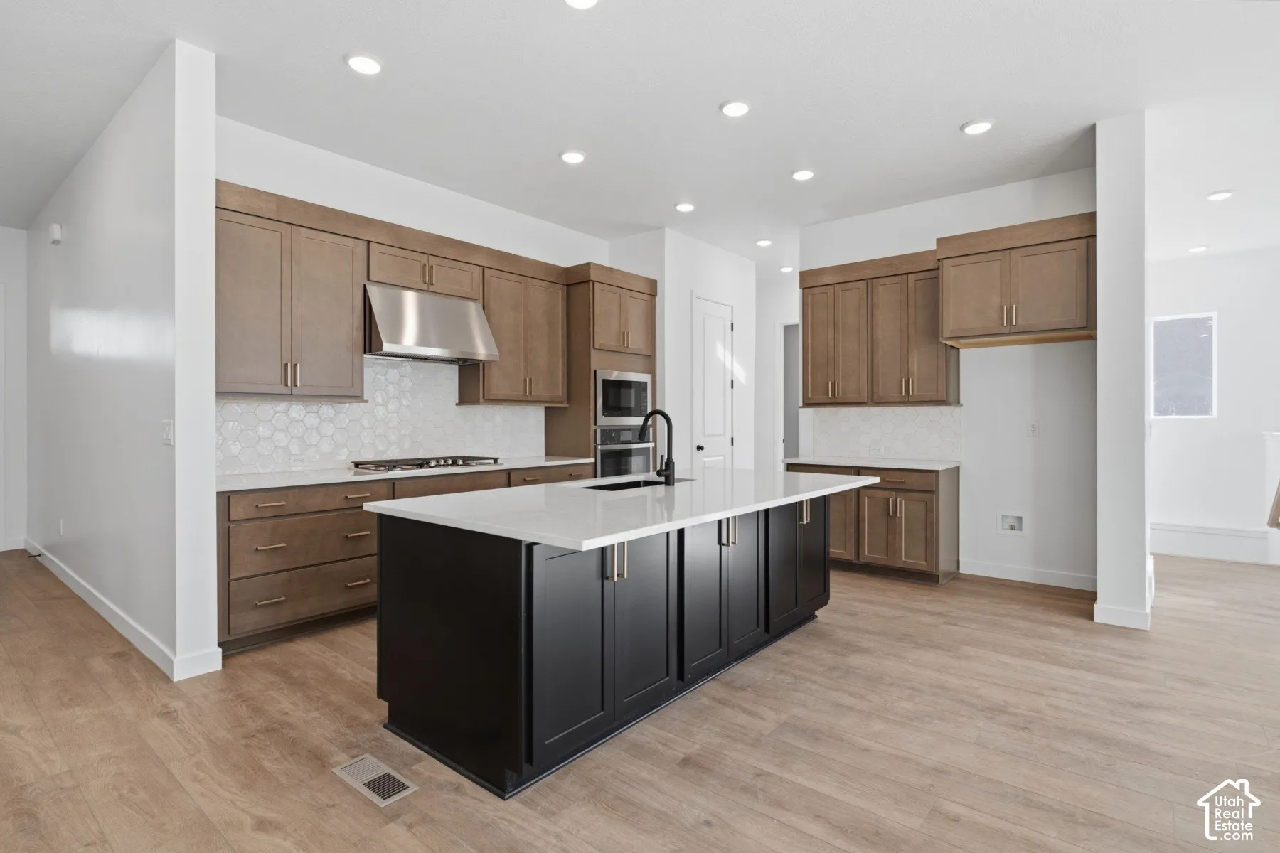Kitchen featuring wood-finish flooring, a central island with a contrasting dark finish, light-toned quartz countertops, built-in stainless steel appliances, and a tiled backsplash