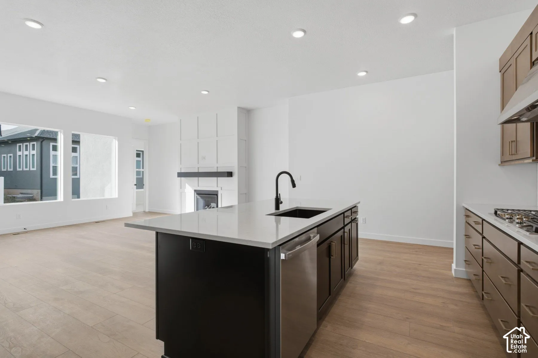 Kitchen featuring wood-finish cabinetry, stone-surface island, undermount sink with matte black faucet, and built-in stainless steel appliances, accented by a wall-mounted fireplace with custom grid molding and recessed lighting