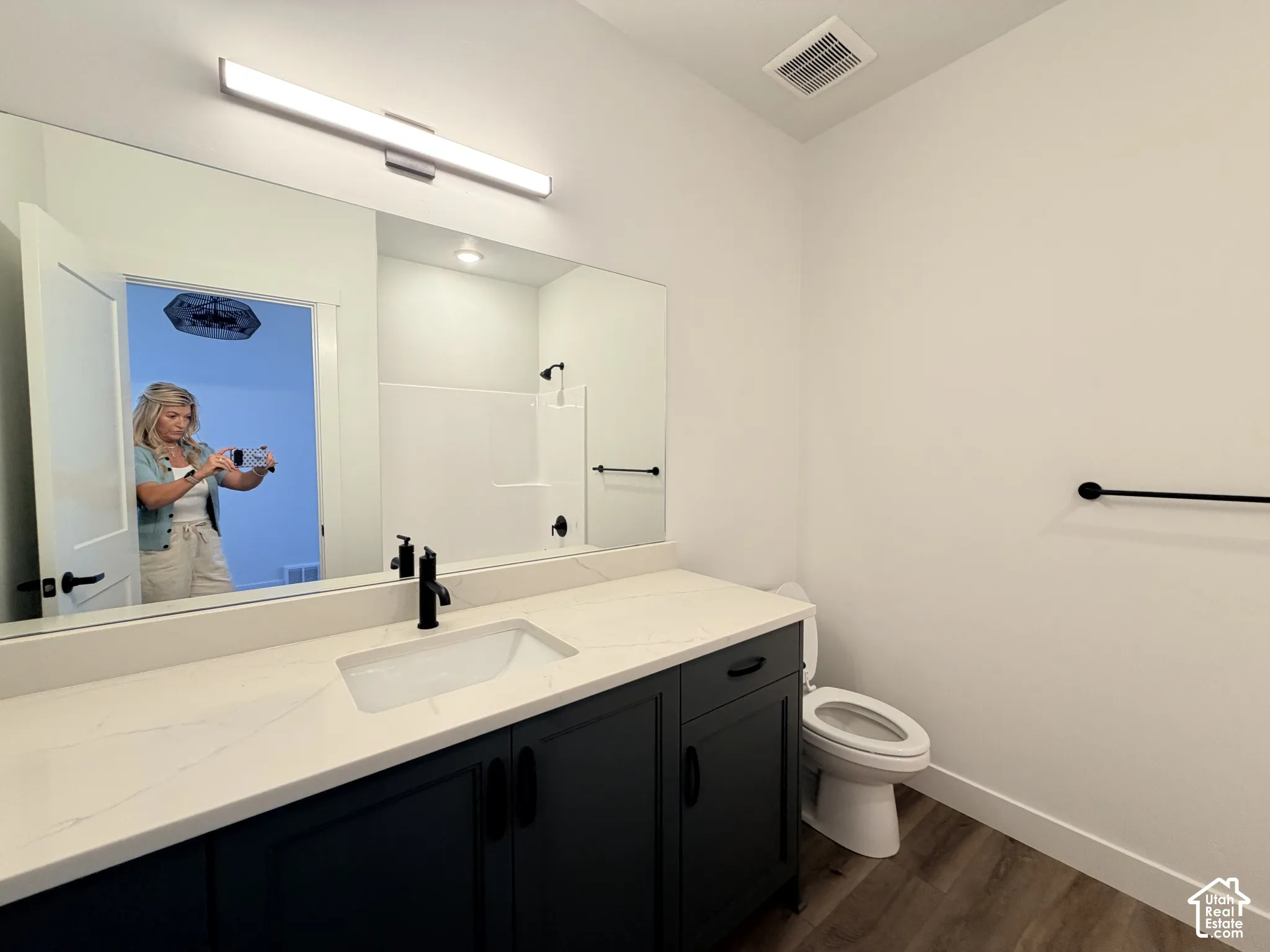 Bathroom featuring a vanity with a white countertop and rectangular undermount sink, dark cabinetry, full-width mirror, matte black fixtures, and wood-finish flooring