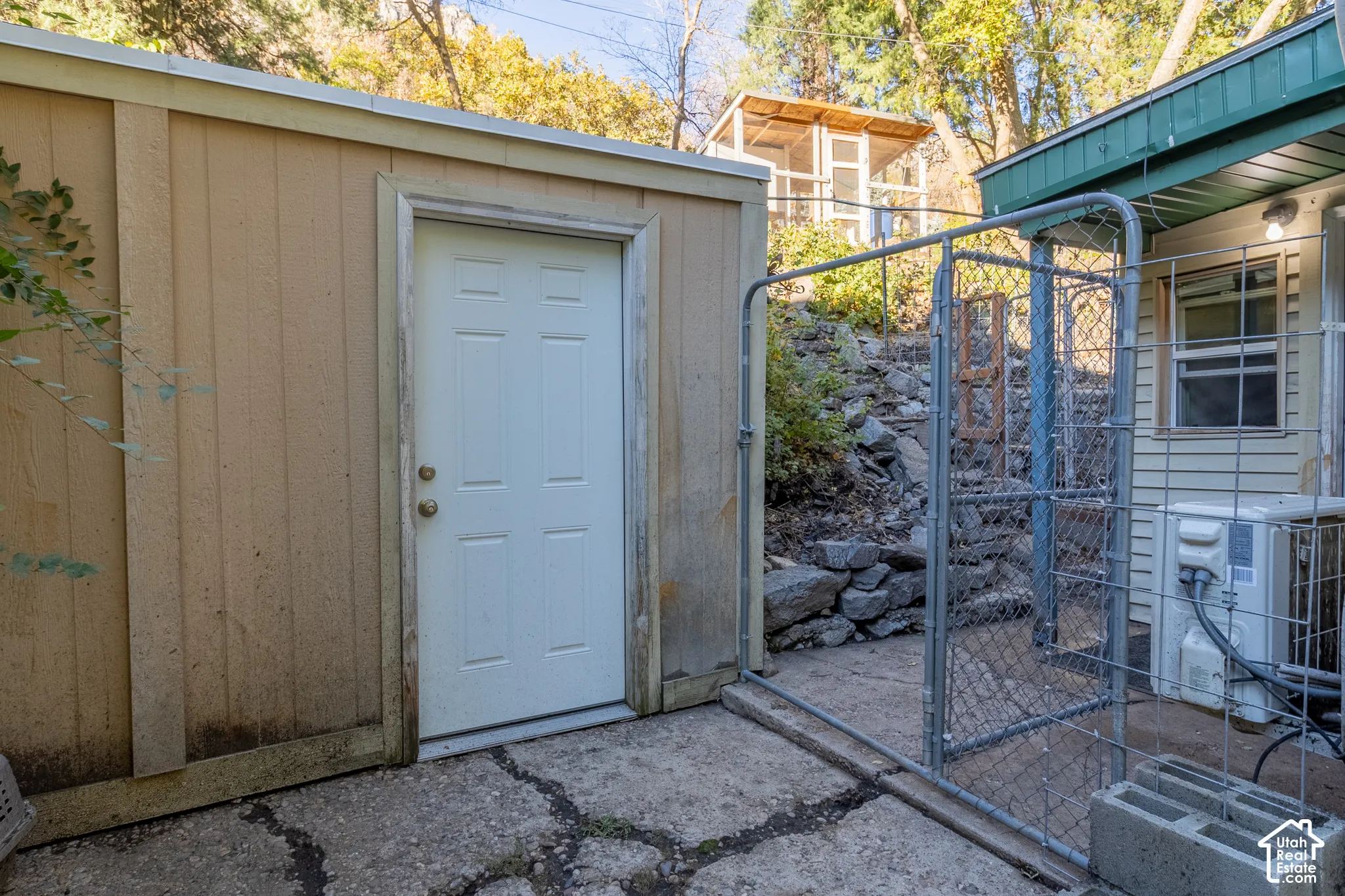 Exterior storage shed featuring a solid panel door, concrete patio, stone retaining wall, and metal chain-link fencing
