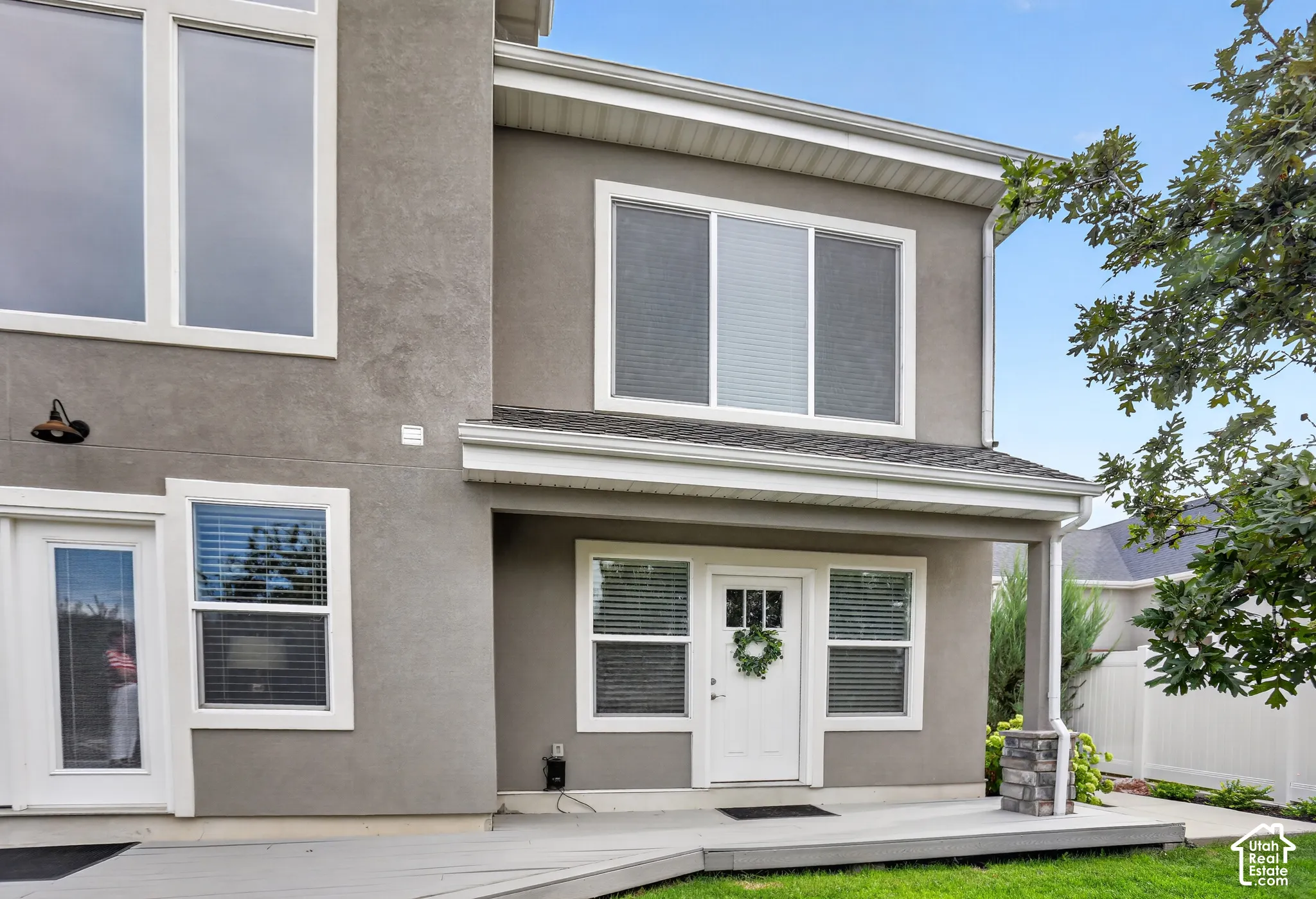 Two-story exterior featuring a neutral stucco finish, multi-pane windows, a covered porch with a stone-clad pillar, and a wood-decked patio