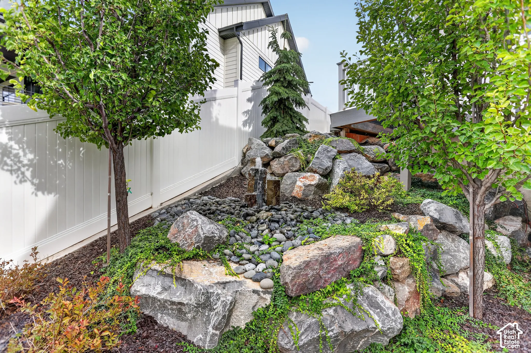 Landscaped backyard featuring a tiered rock garden, integrated stone water feature, mature deciduous trees, and white vinyl privacy fencing