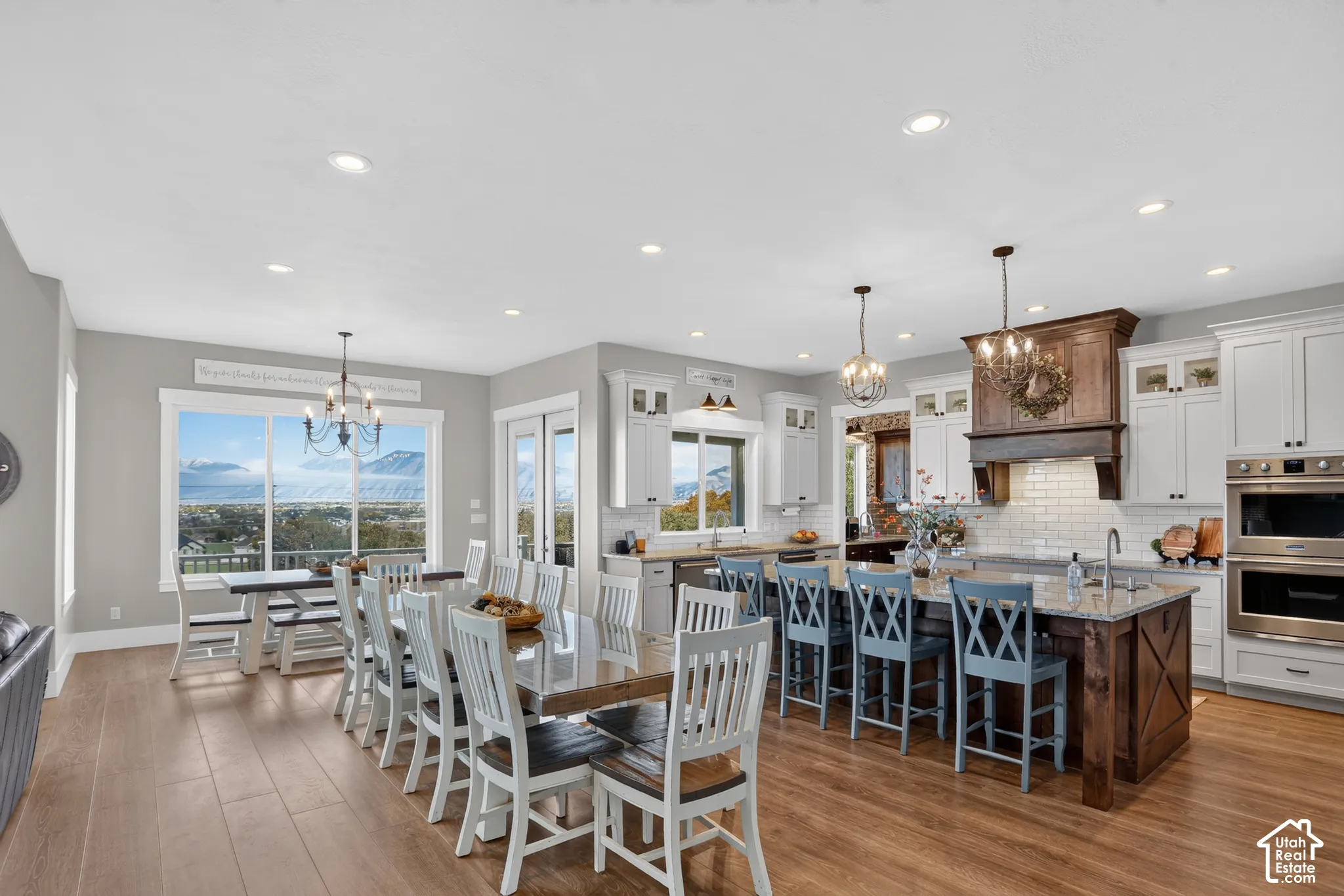 Open-concept kitchen featuring an expansive center island with stone countertops, custom cabinetry with a prominent wooden range hood, subway tile backsplash, recessed lighting, and wood-finish flooring