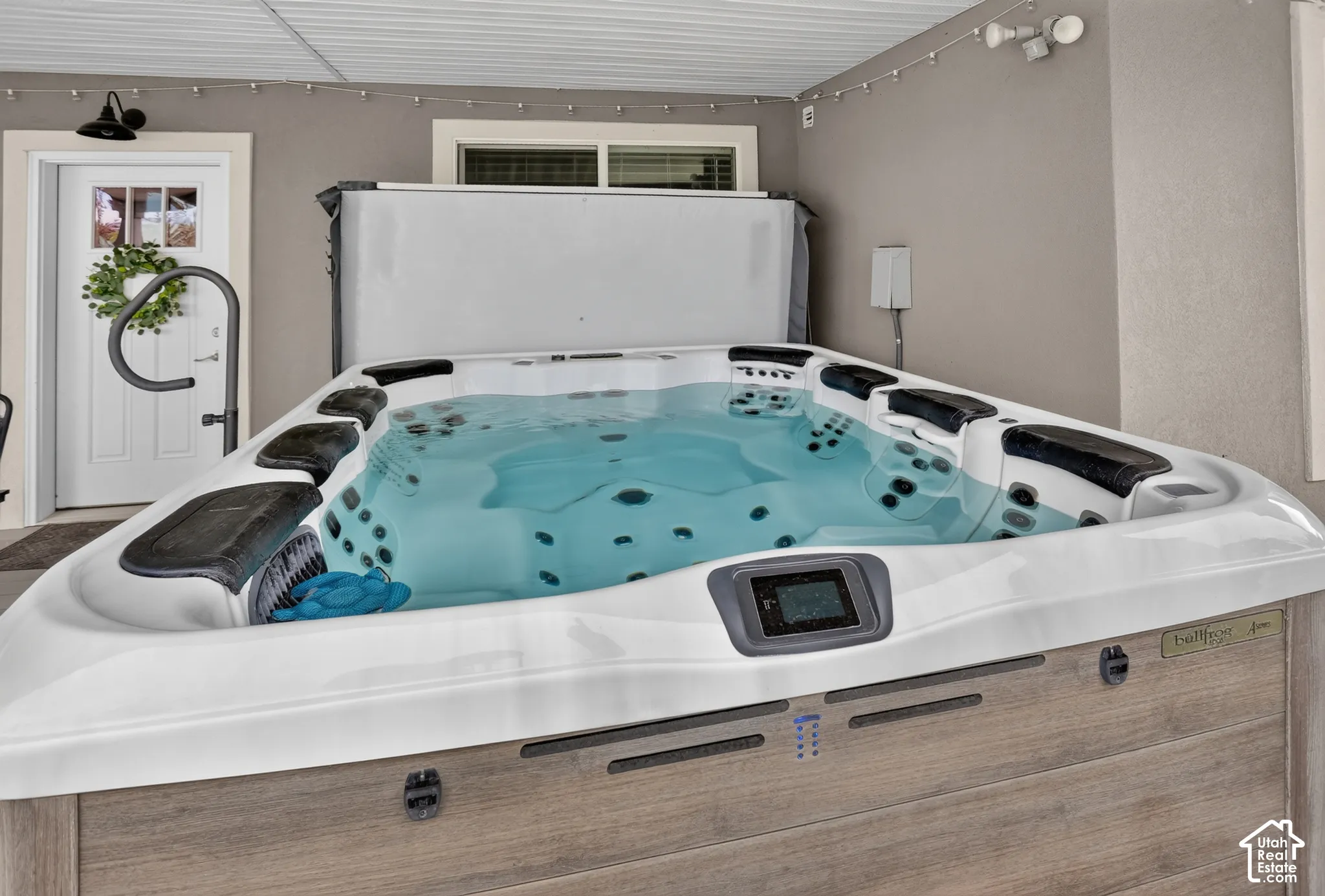 Covered patio featuring an integrated hot tub with headrests and digital control panel, complemented by a white exterior door with glass detailing and wall-mounted exterior lighting
