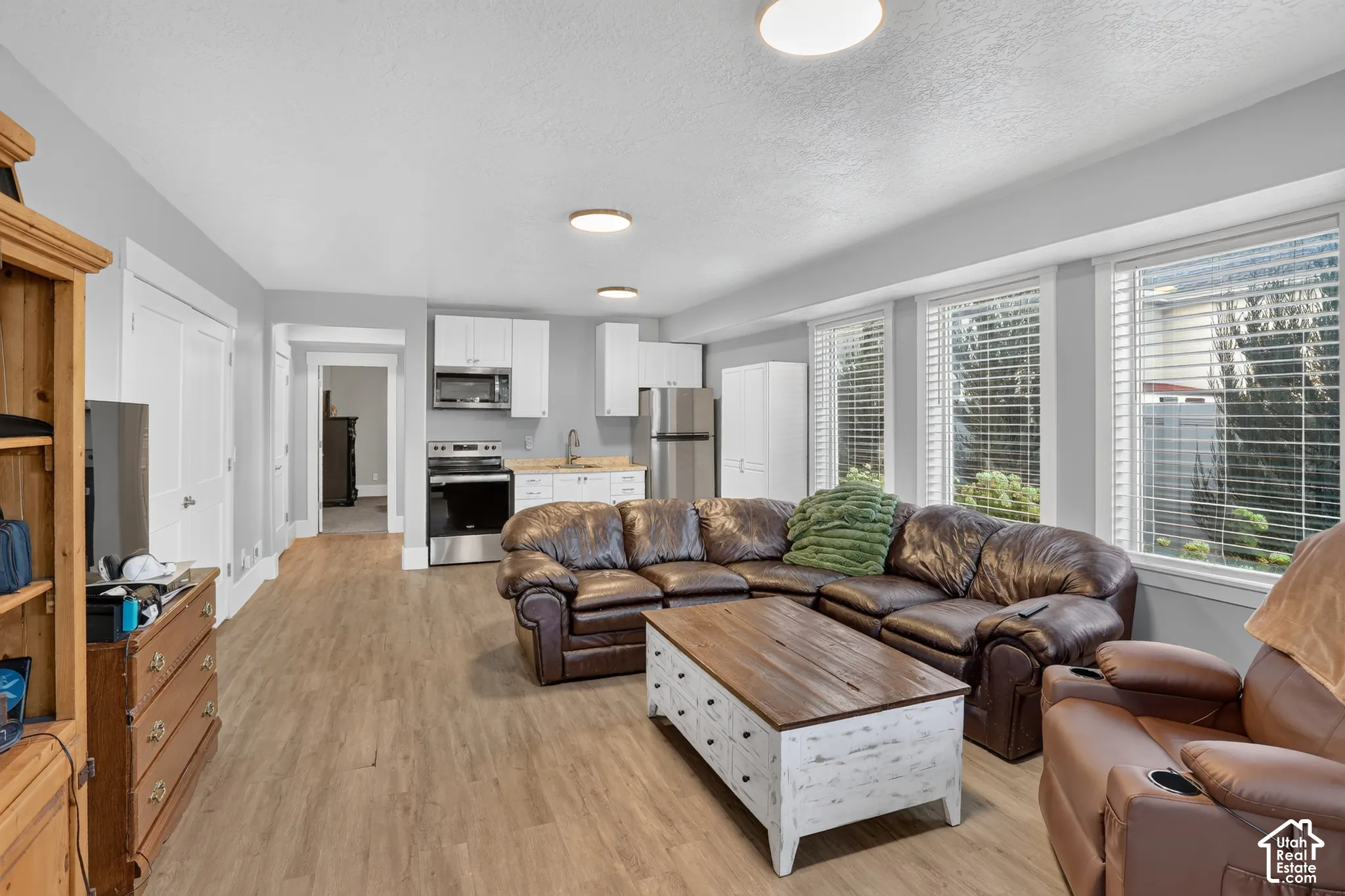 Open-concept living space featuring wood-finish flooring, white cabinetry, stainless steel appliances, neutral wall tones, and large windows with horizontal blinds