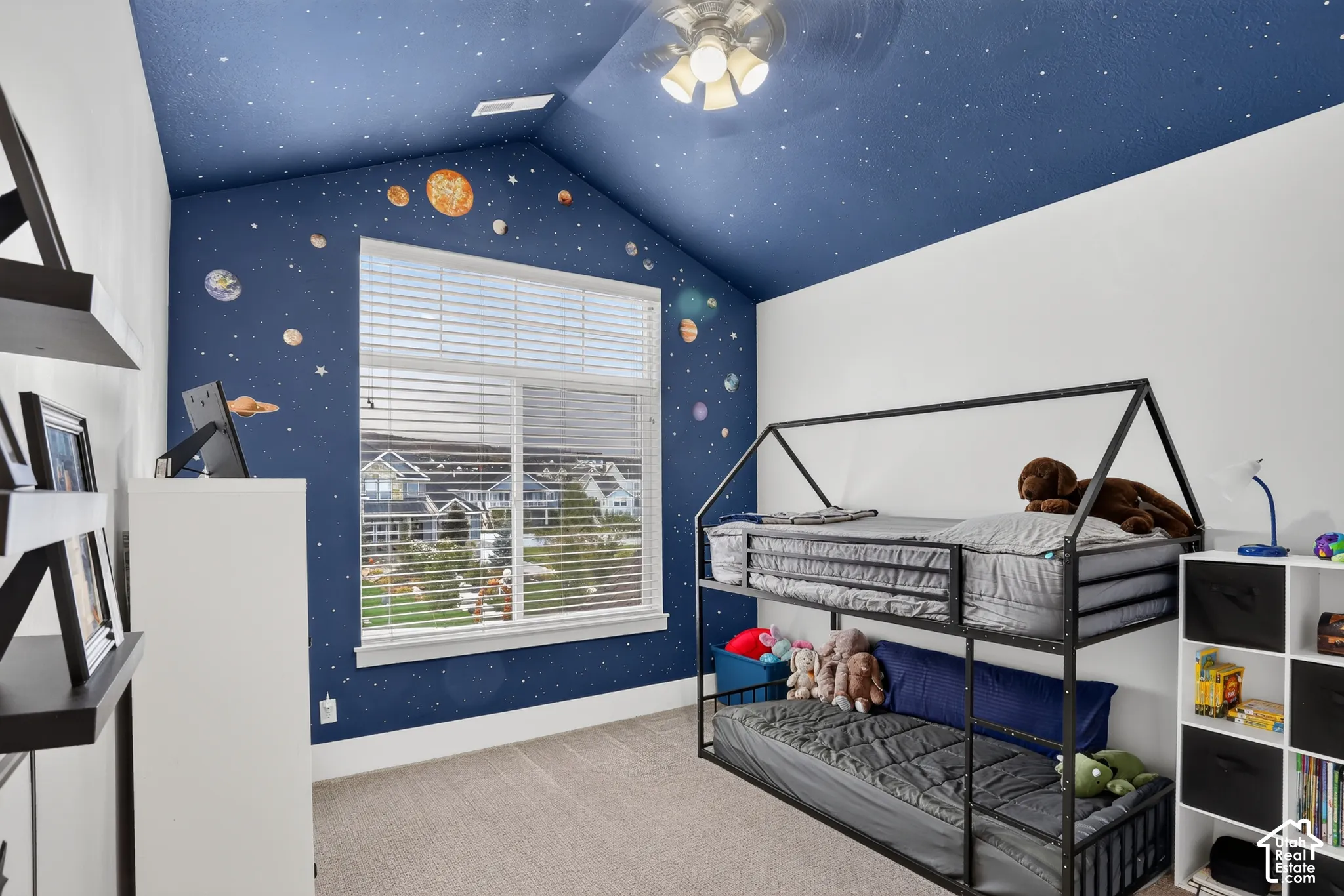 Vaulted ceiling with a custom painted mural, large picture window with horizontal blinds, ceiling fan with integrated lighting, and neutral wall-to-wall carpeting