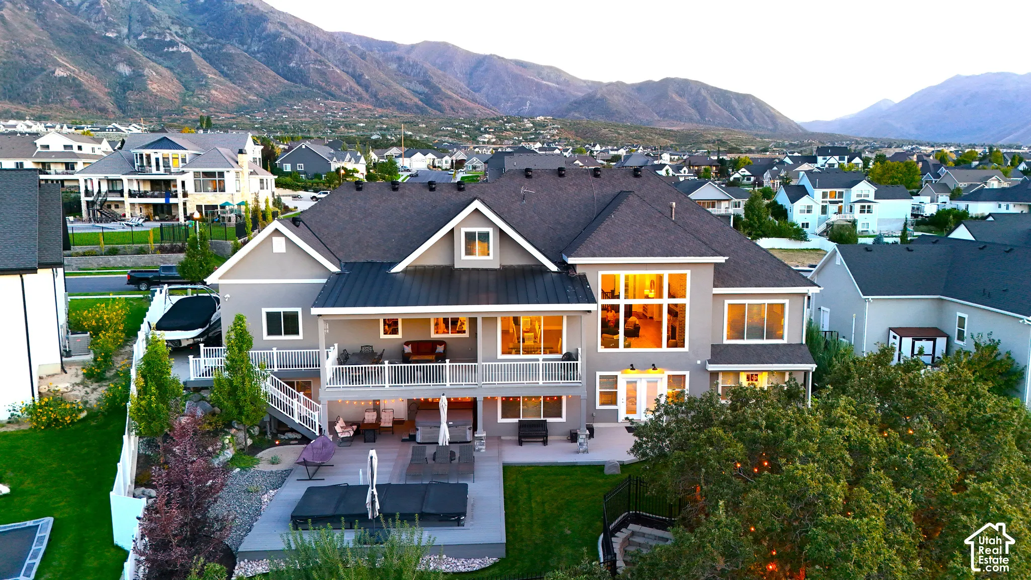 Two-story residence featuring a multi-level deck with white railings, a lower-level concrete patio, large multi-pane windows, and a metal-accented roofline against a mountain backdrop