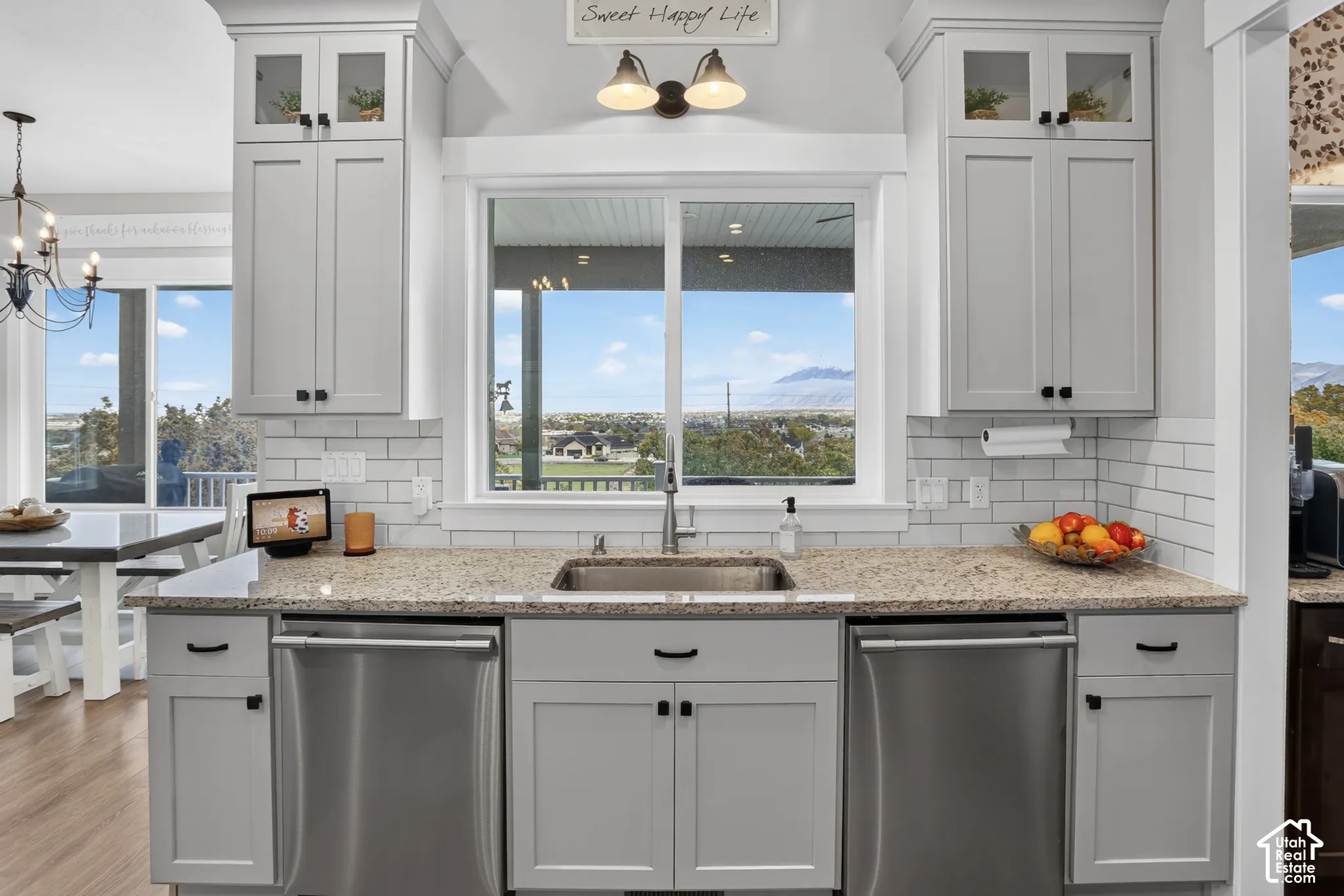 Kitchen featuring light-toned cabinetry with glass-front uppers, stone-finish countertops, stainless steel dual dishwashers, subway tile backsplash, and a large central window with mountain scenery