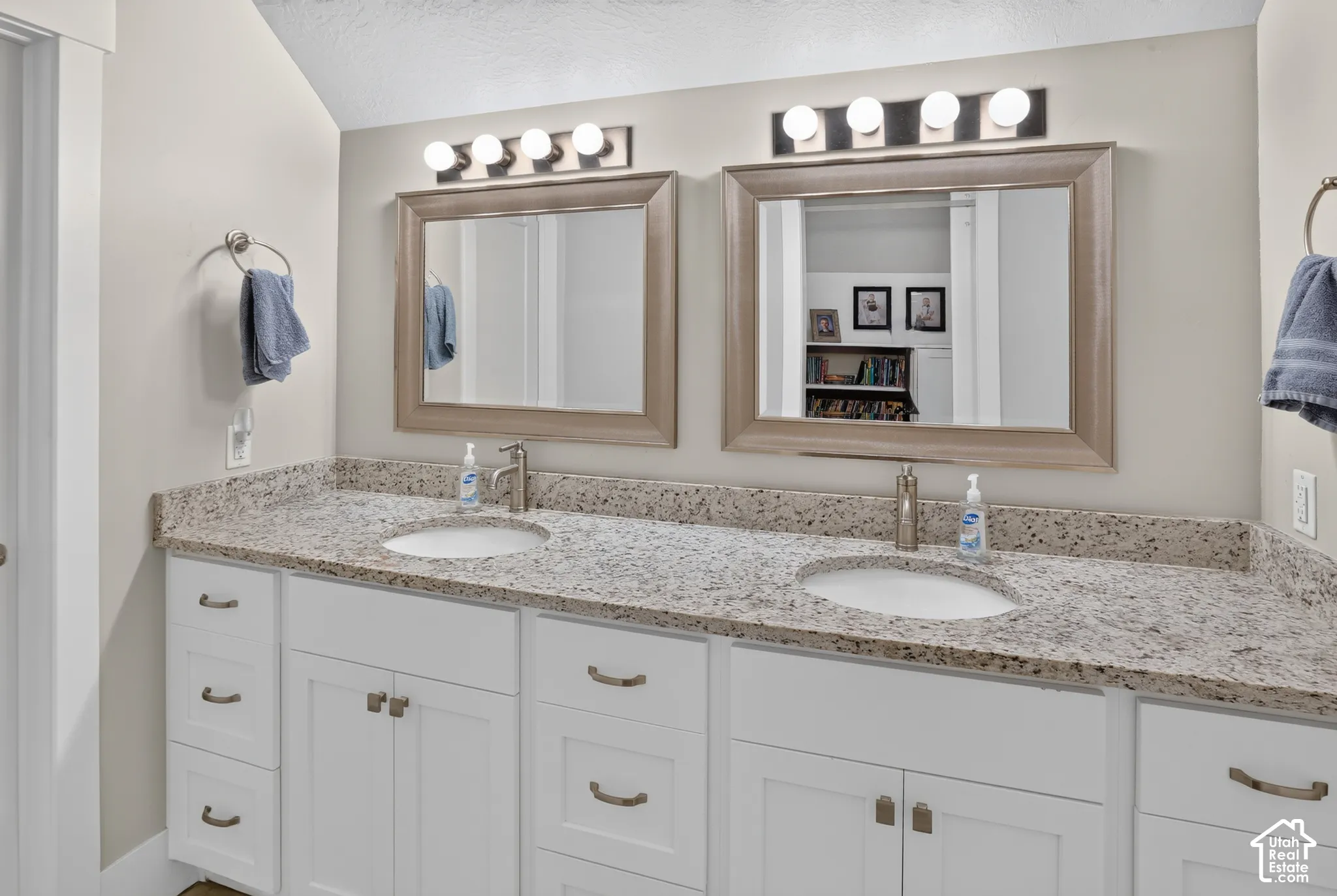 Dual-sink vanity with stone-finish countertop, white Shaker-style cabinetry, two framed mirrors, and dual four-light wall-mounted fixtures