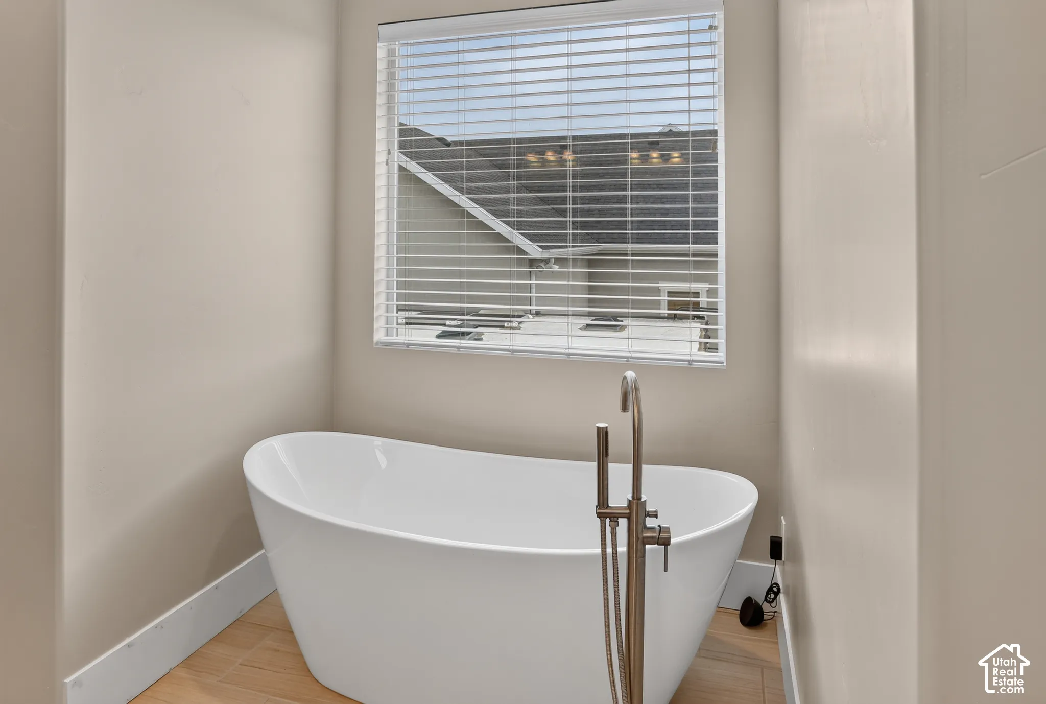 Freestanding white soaking tub with a floor-mounted brushed metal faucet, set against wood-finish flooring and a window with horizontal blinds