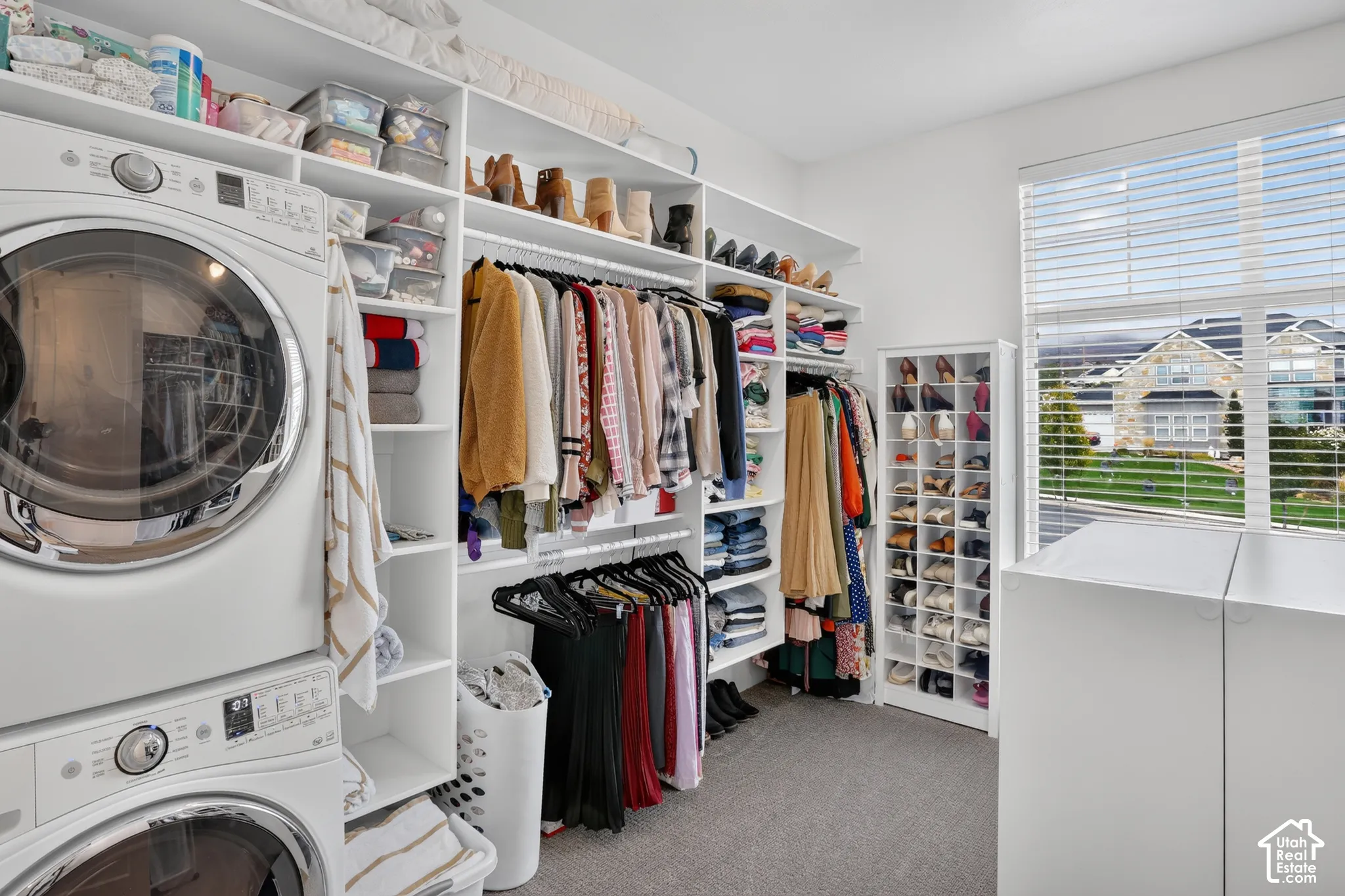 Built-in white shelving system featuring custom garment rods, tiered shoe cubbies, integrated laundry appliances, and large window with horizontal blinds