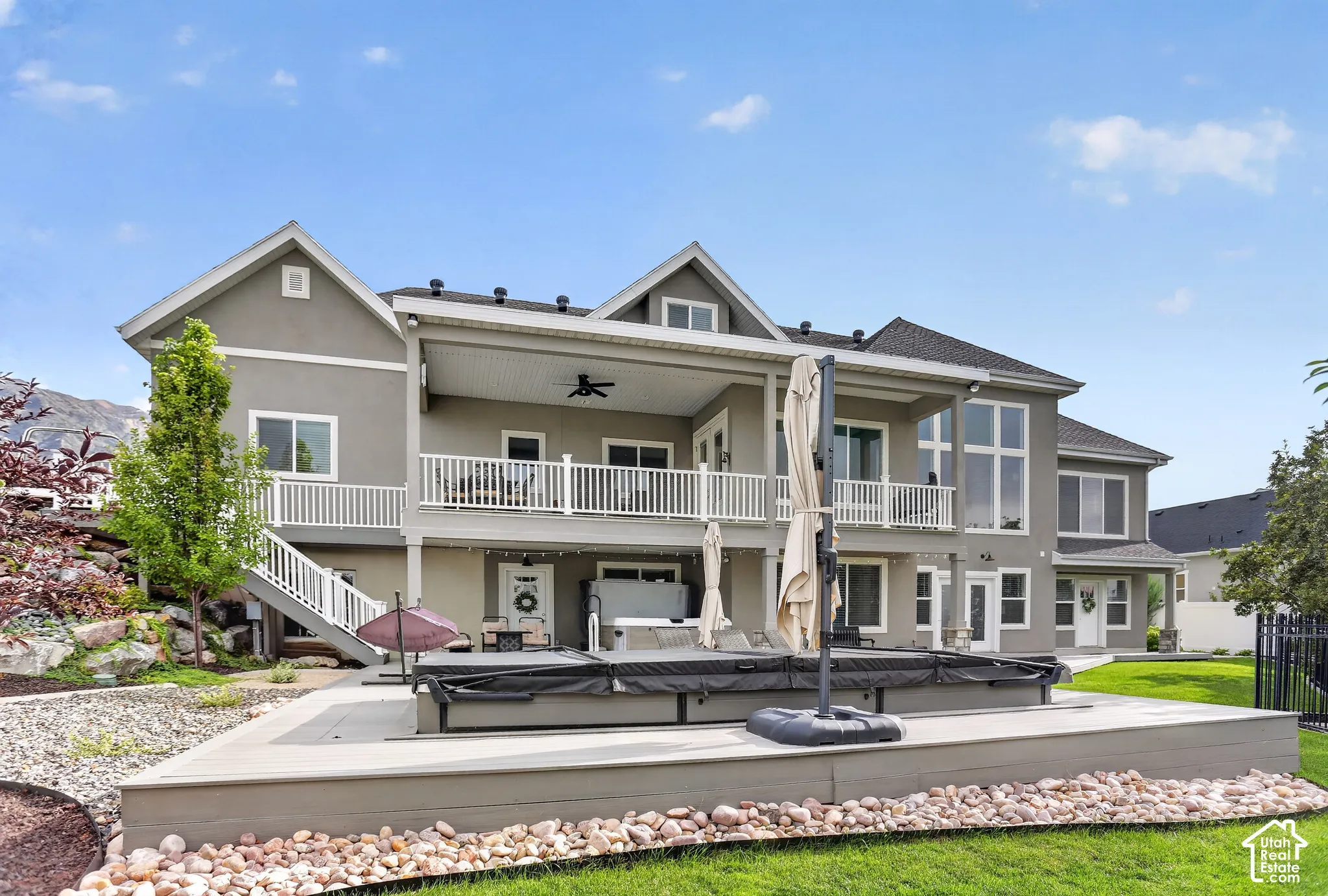 Multi-level rear exterior featuring a covered deck with white railing, a ground-level patio, built-in hot tub, and mountain backdrop