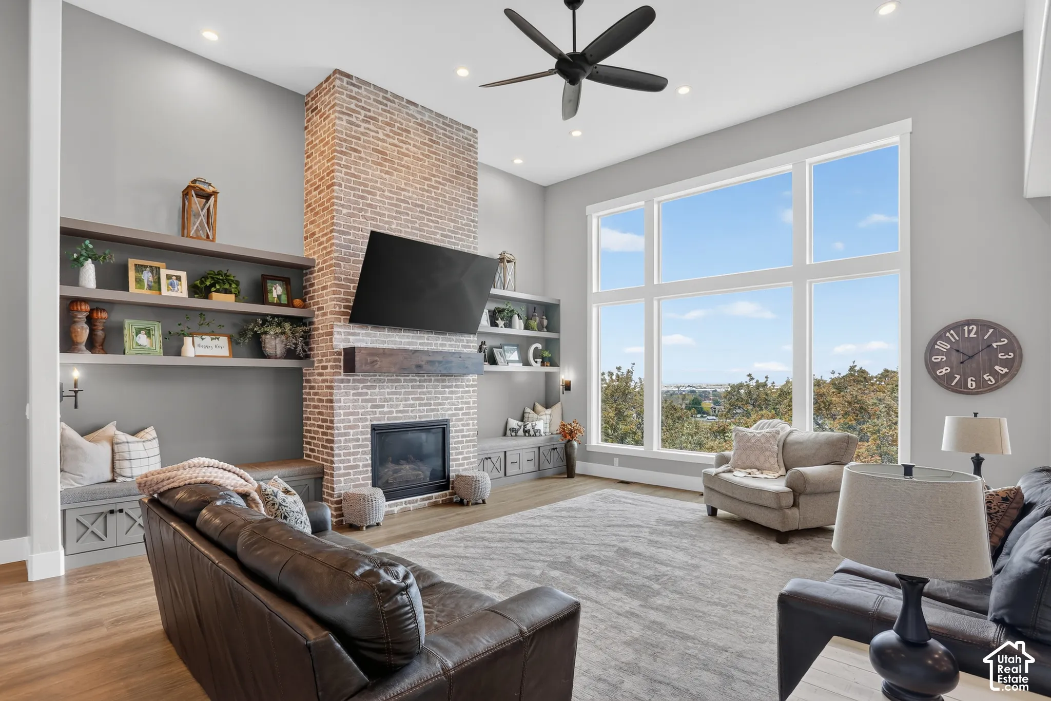 High-ceilinged living area featuring a floor-to-ceiling brick fireplace, natural wood-finish flooring, built-in cabinetry with floating shelving, and expansive grid windows