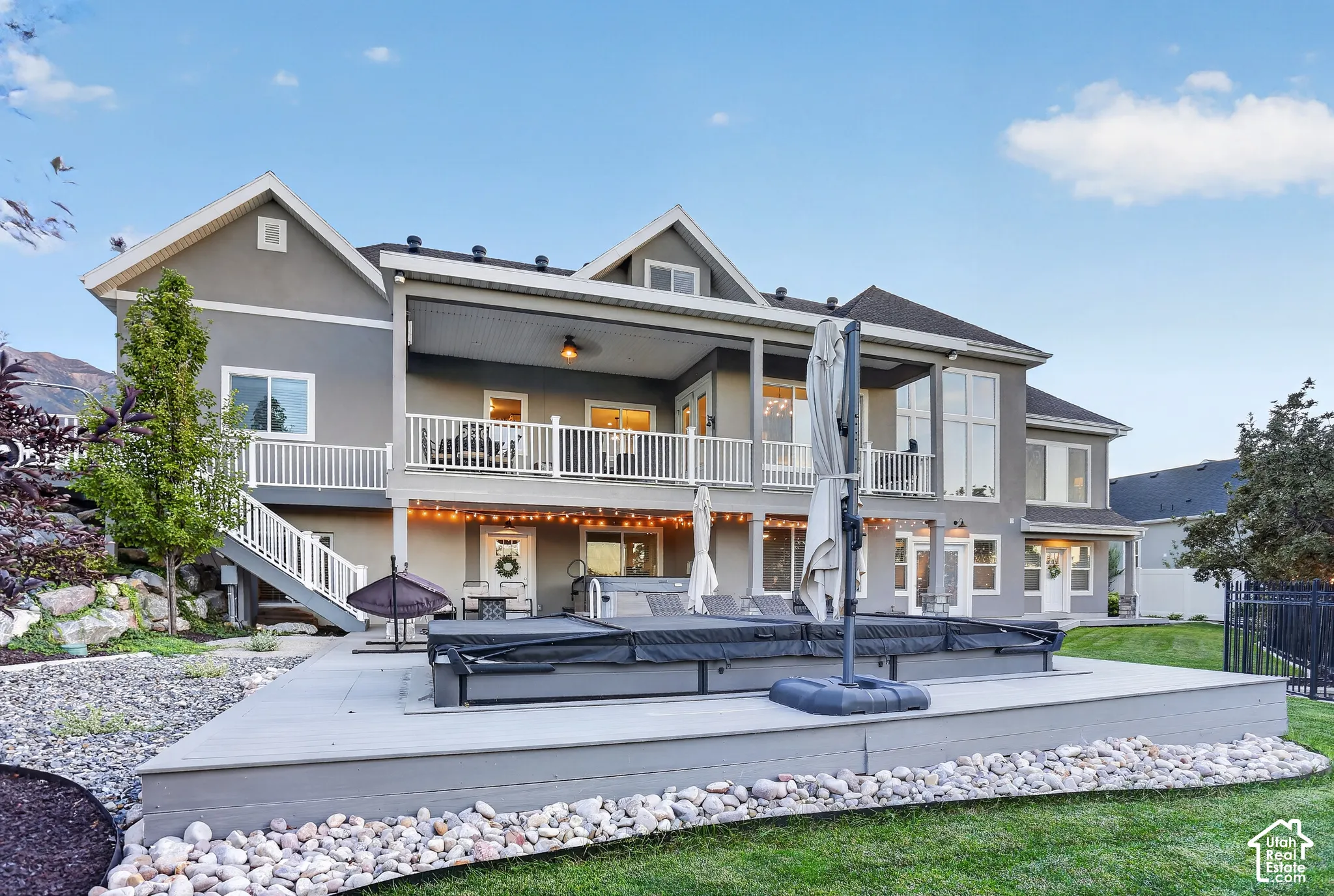 Multi-level exterior featuring a covered deck with a ceiling fan, a lower-level concrete patio, a built-in spa, white metal railings, and stone-accented landscape borders