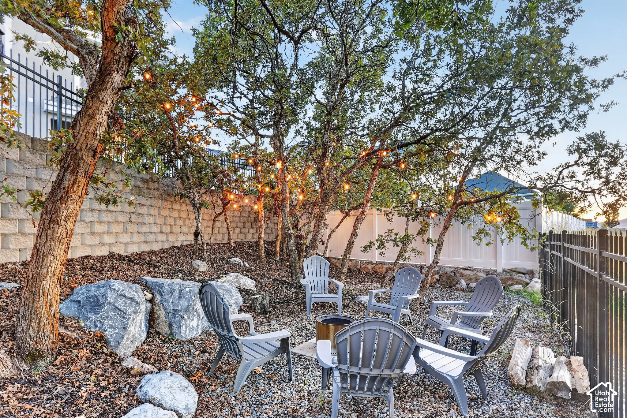 Backyard area featuring a gravel ground surface, mature trees, concrete block retaining wall, and perimeter metal and vinyl fencing