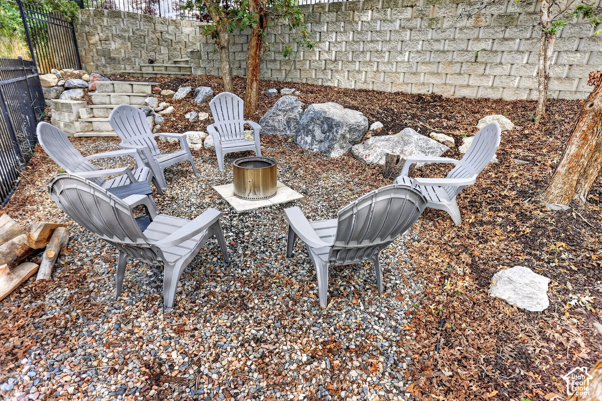 Gravel-surfaced fire pit area featuring a metal fire cylinder, stacked stone retaining wall, decorative boulders, and tiered stone steps