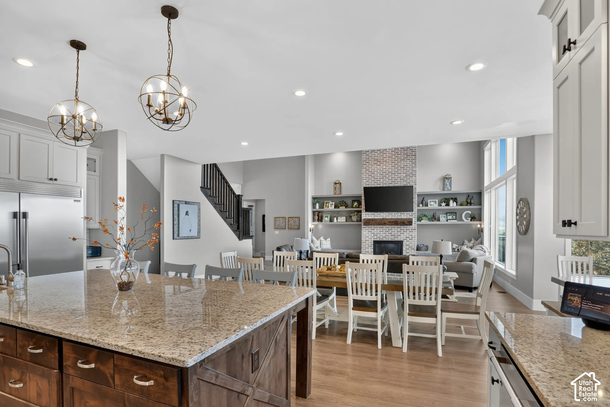 Kitchen island with stone countertop and wood-finish cabinetry, floor-to-ceiling fireplace with brick surround, recessed lighting, spherical pendant fixtures, and tall windows
