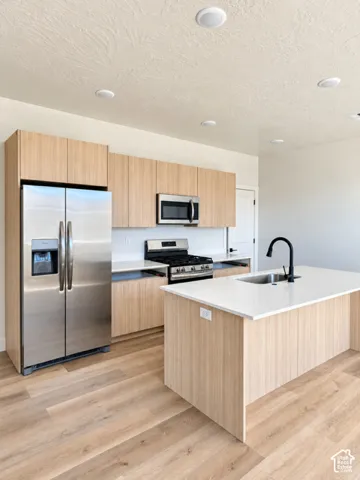 Kitchen featuring flat-panel wood-finish cabinetry, light-toned countertops, stainless steel appliances, a center island with an undermount sink and matte black faucet, and wood-finish flooring