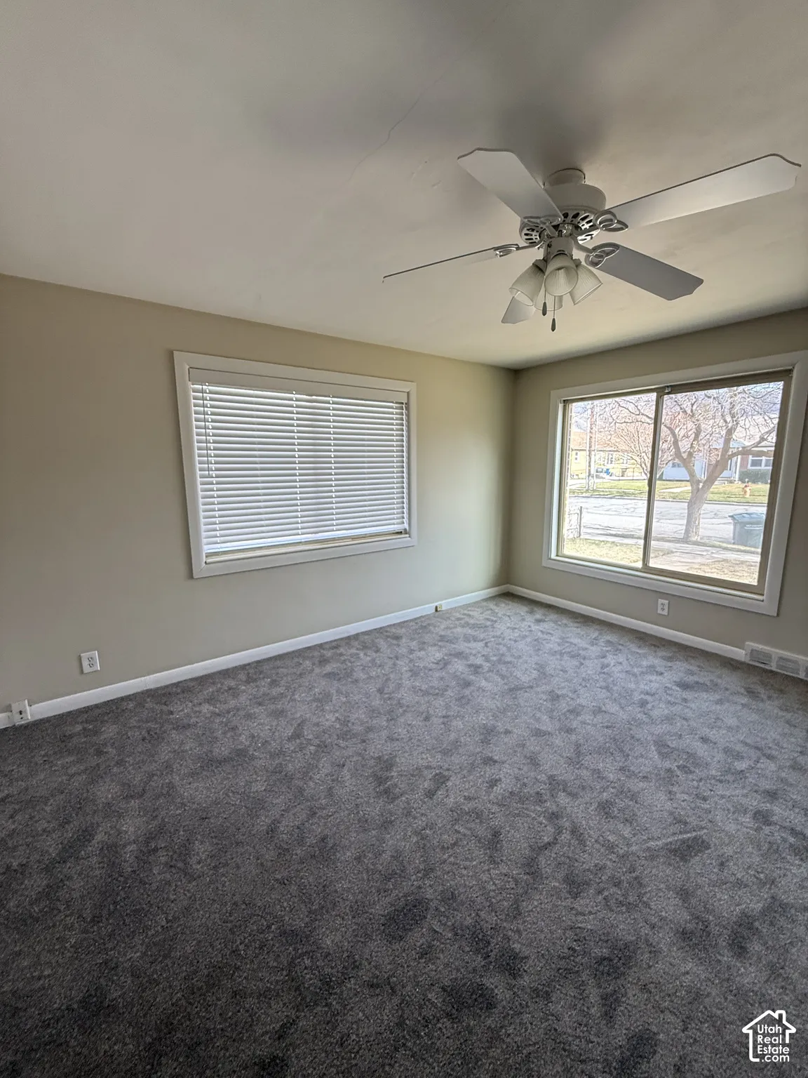 Room interior featuring neutral wall paint, wall-to-wall carpeting, a multi-blade ceiling fan with integrated light fixture, and large windows with horizontal blinds