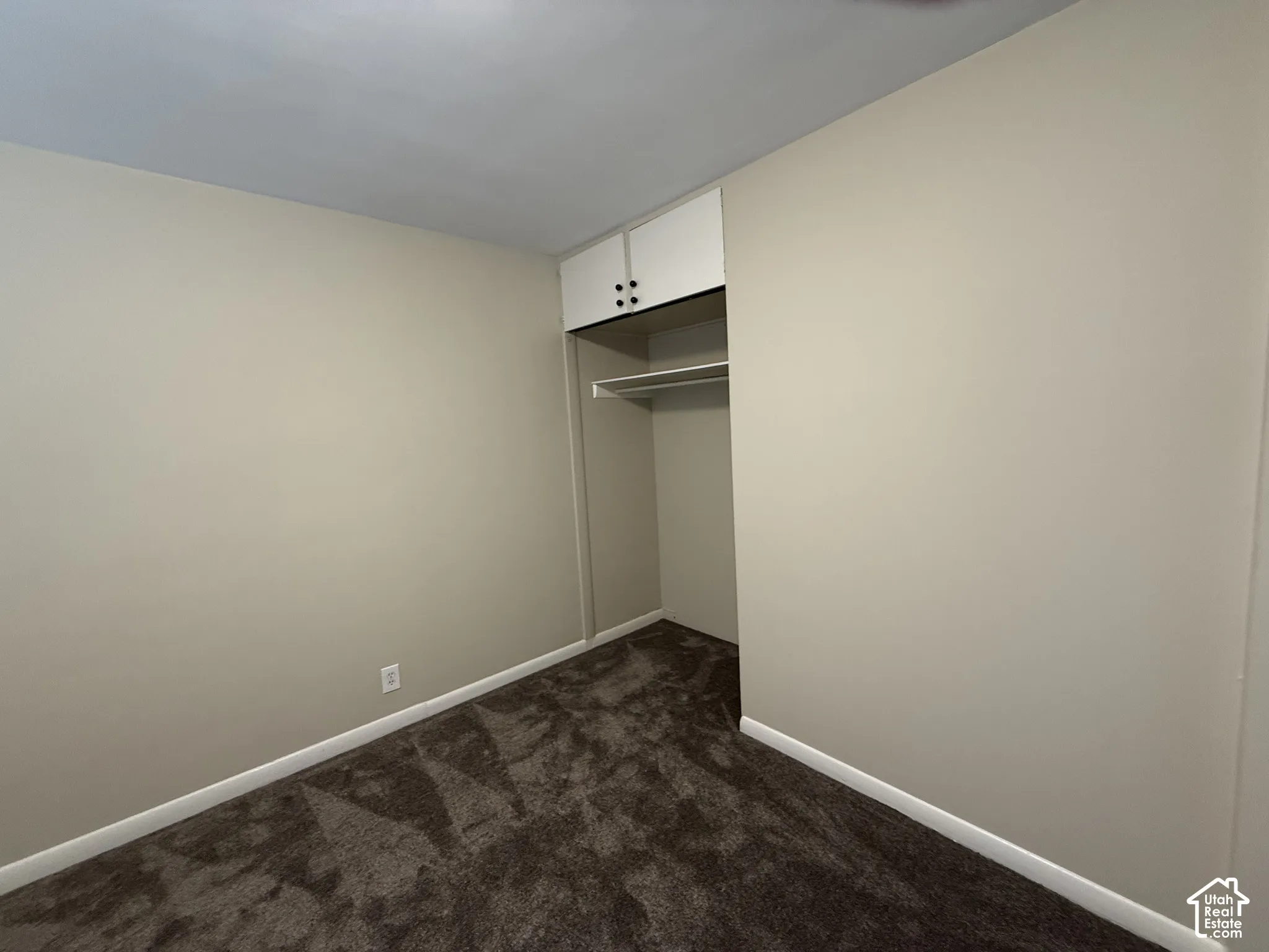 Neutral-toned interior space featuring dark wall-to-wall carpeting and a recessed closet with built-in overhead cabinetry
