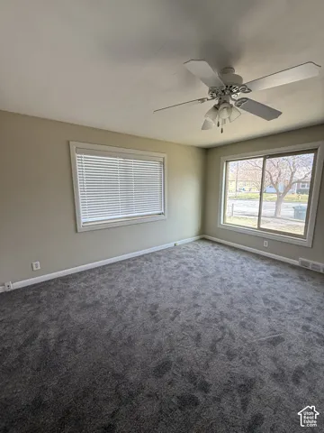 Room interior featuring neutral wall paint, wall-to-wall carpeting, a multi-blade ceiling fan with integrated light fixture, and large windows with horizontal blinds
