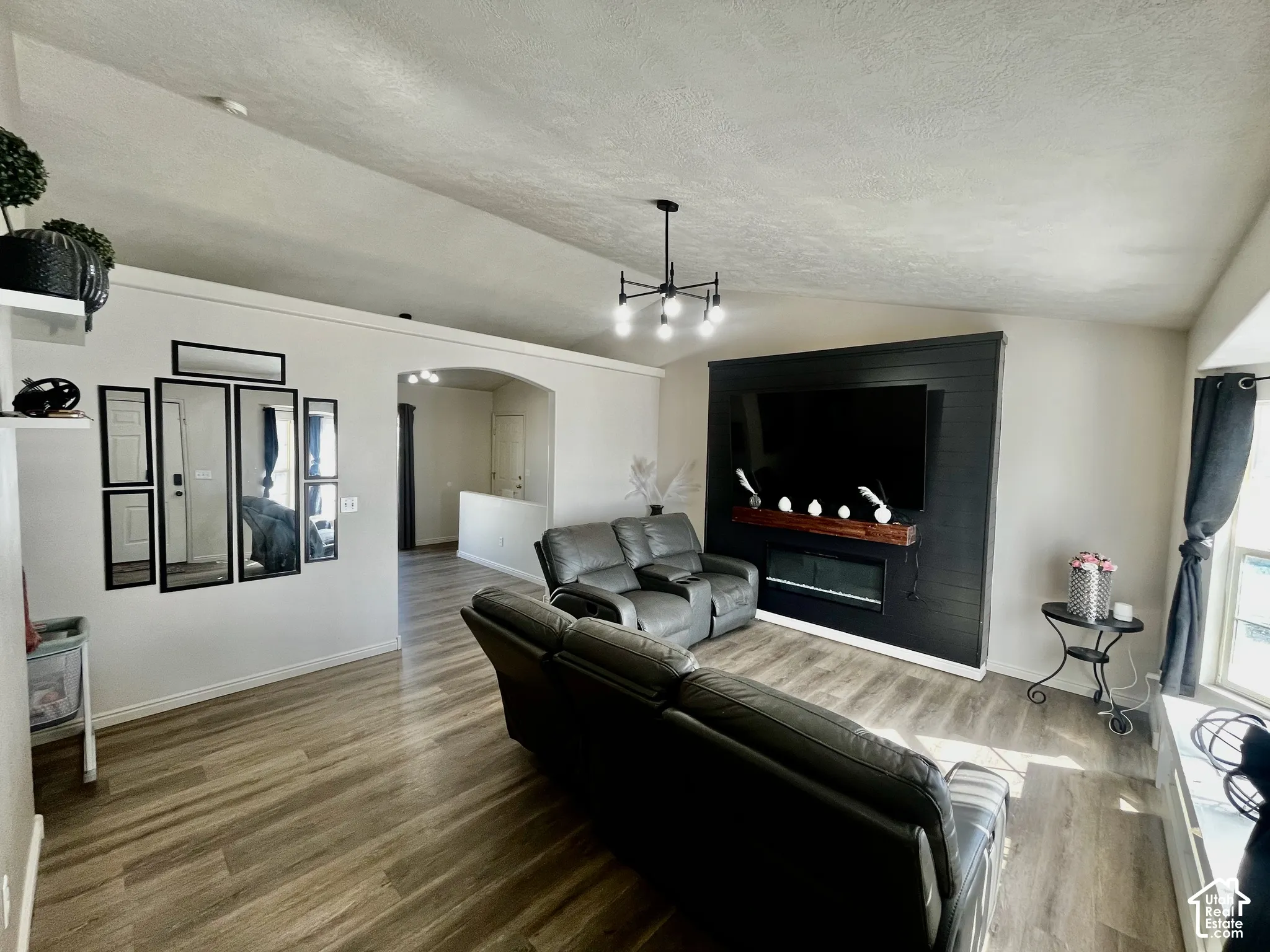 Vaulted ceilings, wood-finish flooring, accent wall with integrated electric fireplace and timber mantel, and multi-arm chandelier