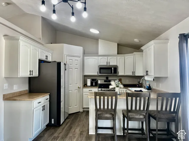 Kitchen featuring white cabinetry with dark hardware, neutral-toned countertops, wood-finish flooring, stainless steel appliances, and a vaulted ceiling with multi-bulb pendant lighting